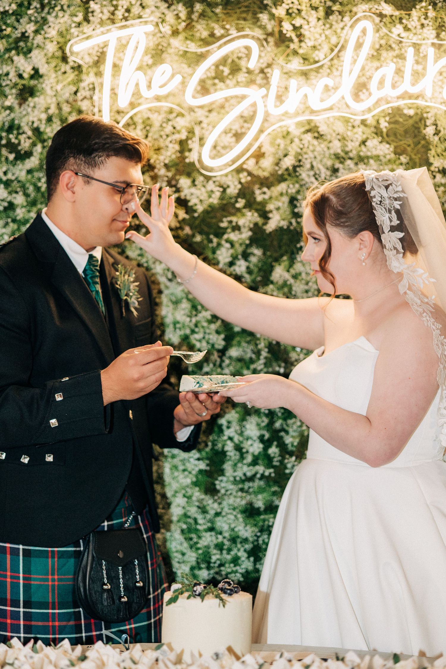 A couple in wedding attire share a sweet cake-feeding moment under a neon sign at their reception.