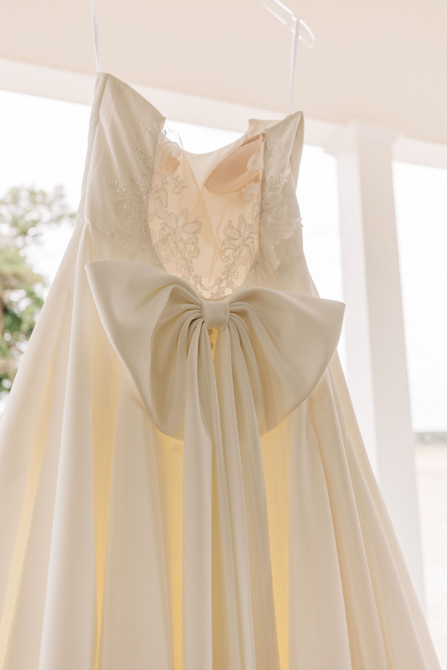 A white wedding dress with lace bodice and large bow detail hanging on display in a light-filled porch.