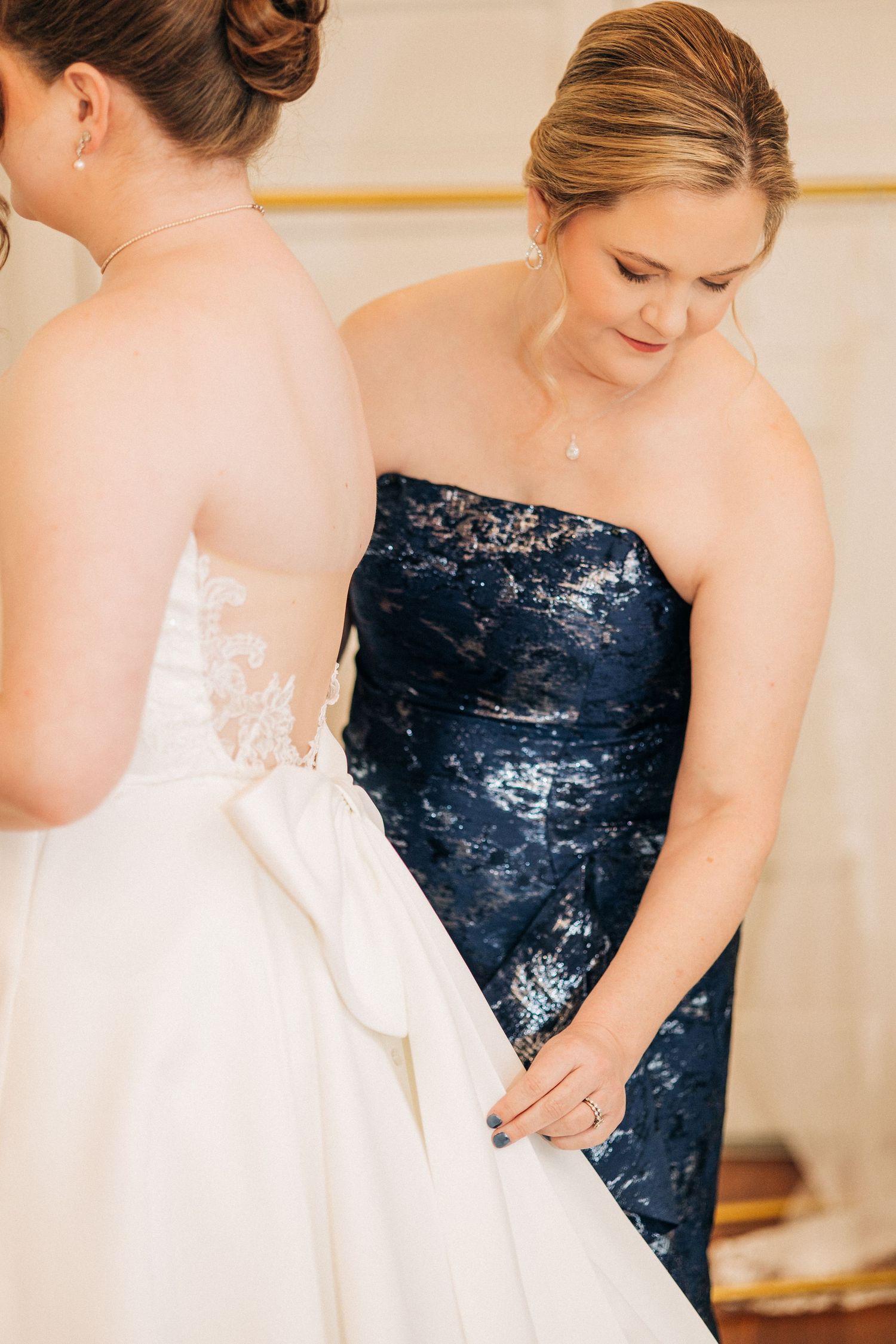 Bridesmaid in a navy sequined gown assists with the bride's white wedding dress.
