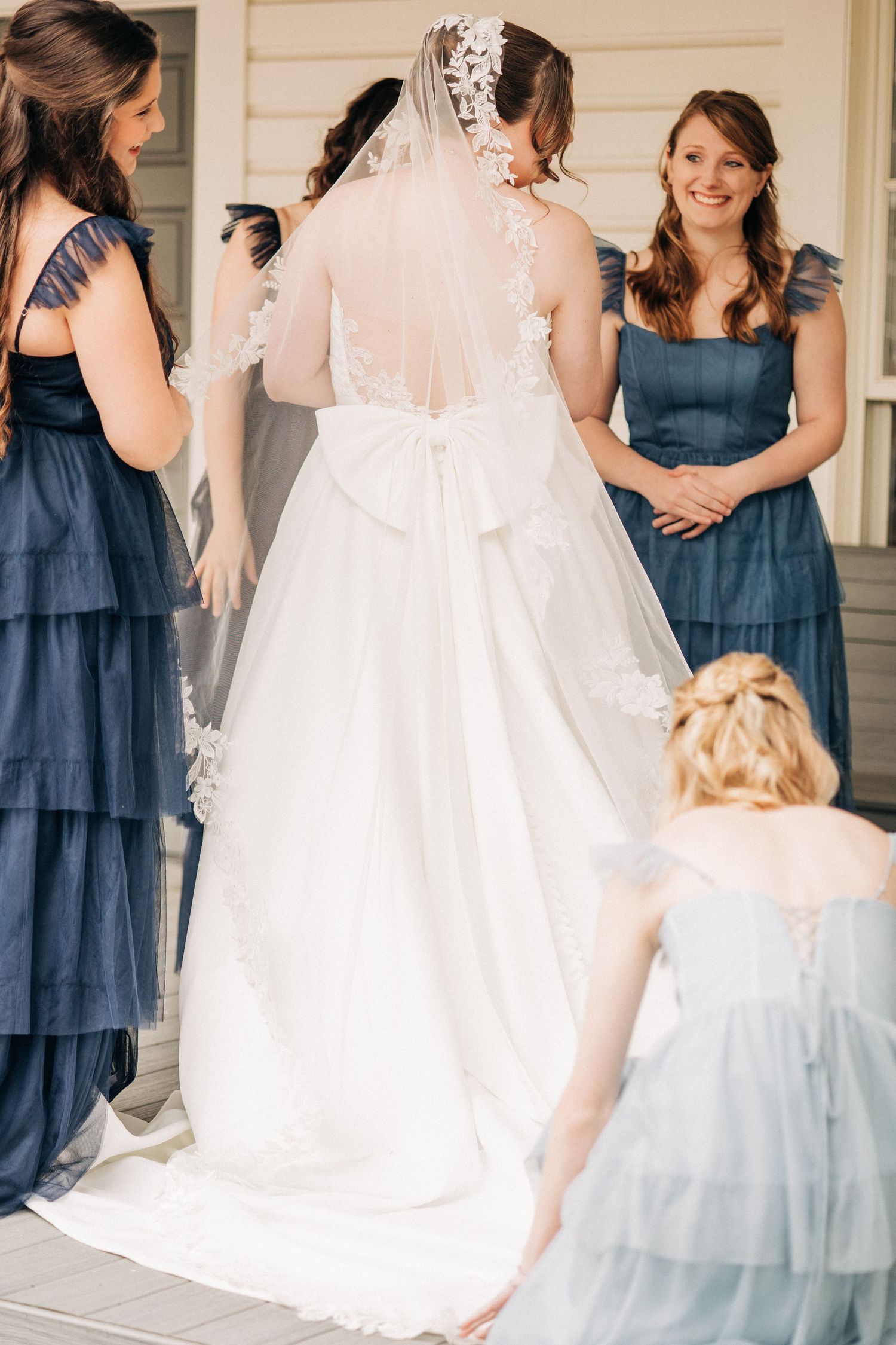 Wedding party poses together on porch wearing navy blue dresses and a flowing white gown with lace details.