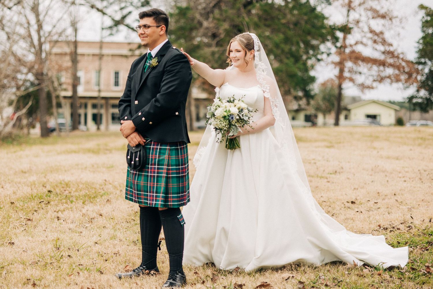 Wedding couple in traditional Scottish dress stand back to back outdoors, with groom in kilt and bride in white gown.