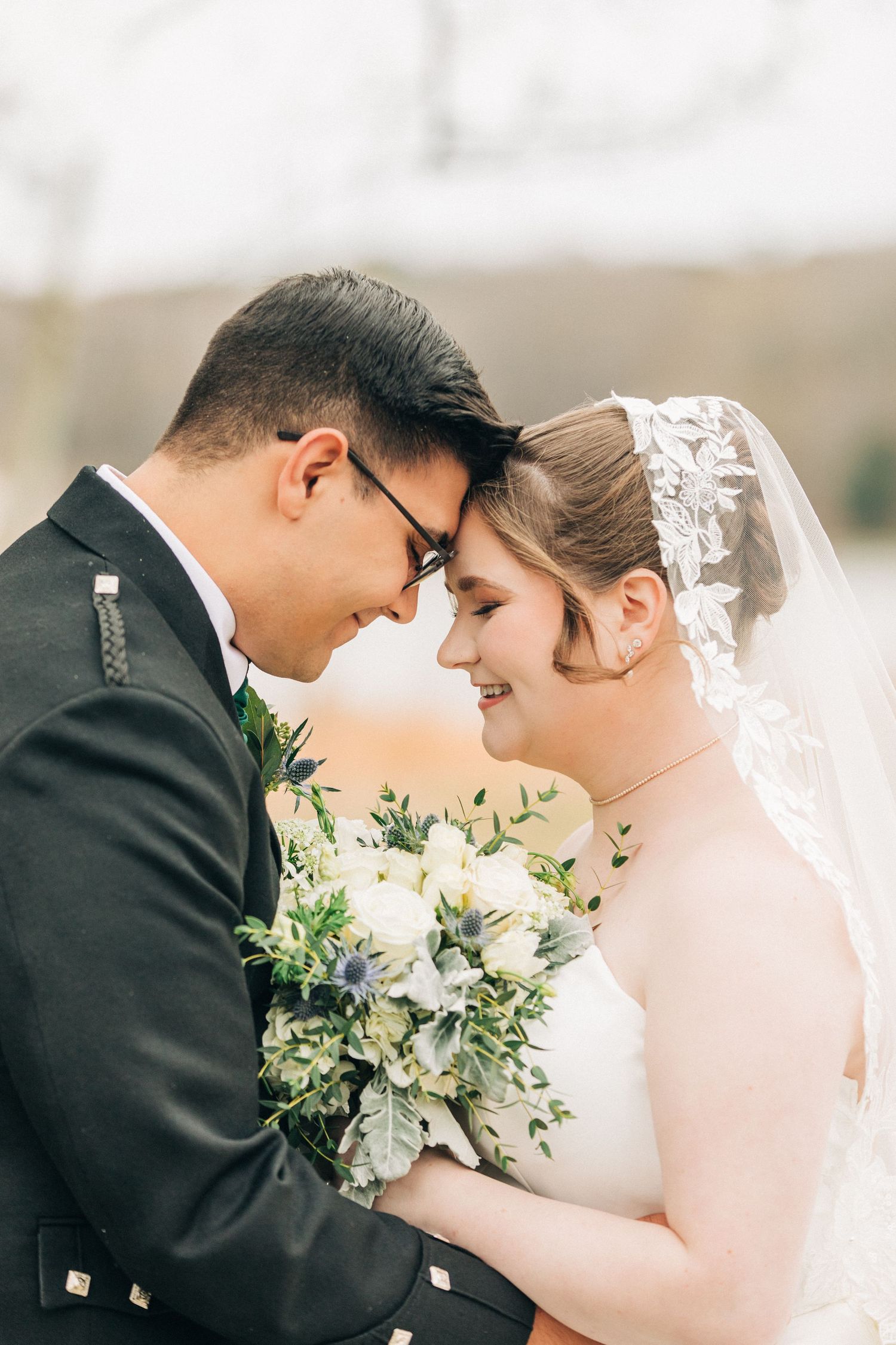 Couple in wedding attire share intimate forehead touch while holding white and green floral bouquet against blurred background.
