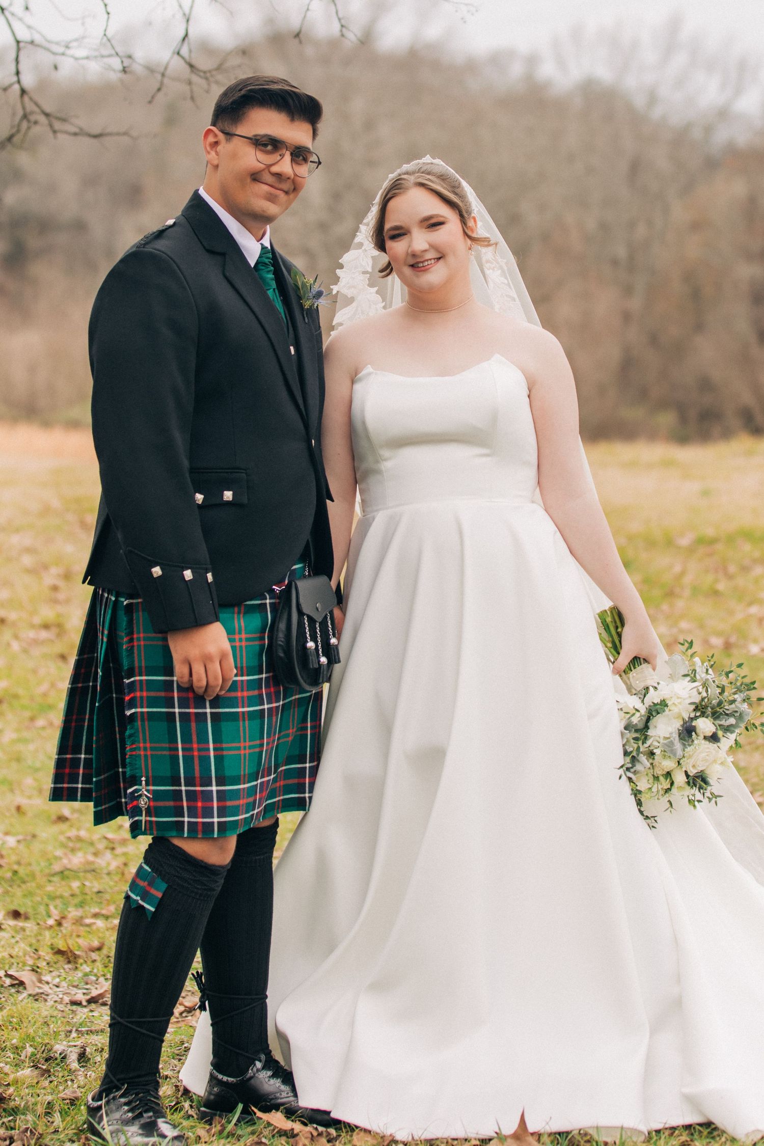 A couple poses outdoors in wedding attire, with the groom wearing a traditional Scottish kilt and the bride in a white gown.