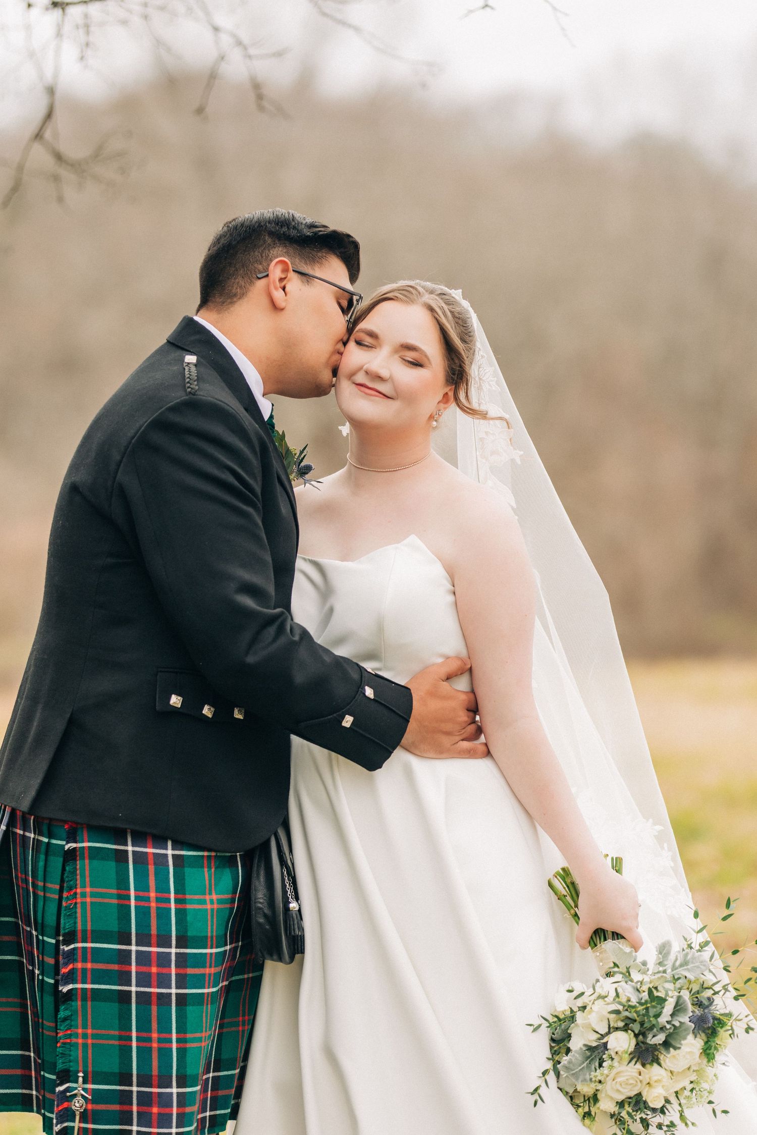 A romantic outdoor wedding moment featuring a groom in traditional Scottish kilt and bride in white dress with bouquet.