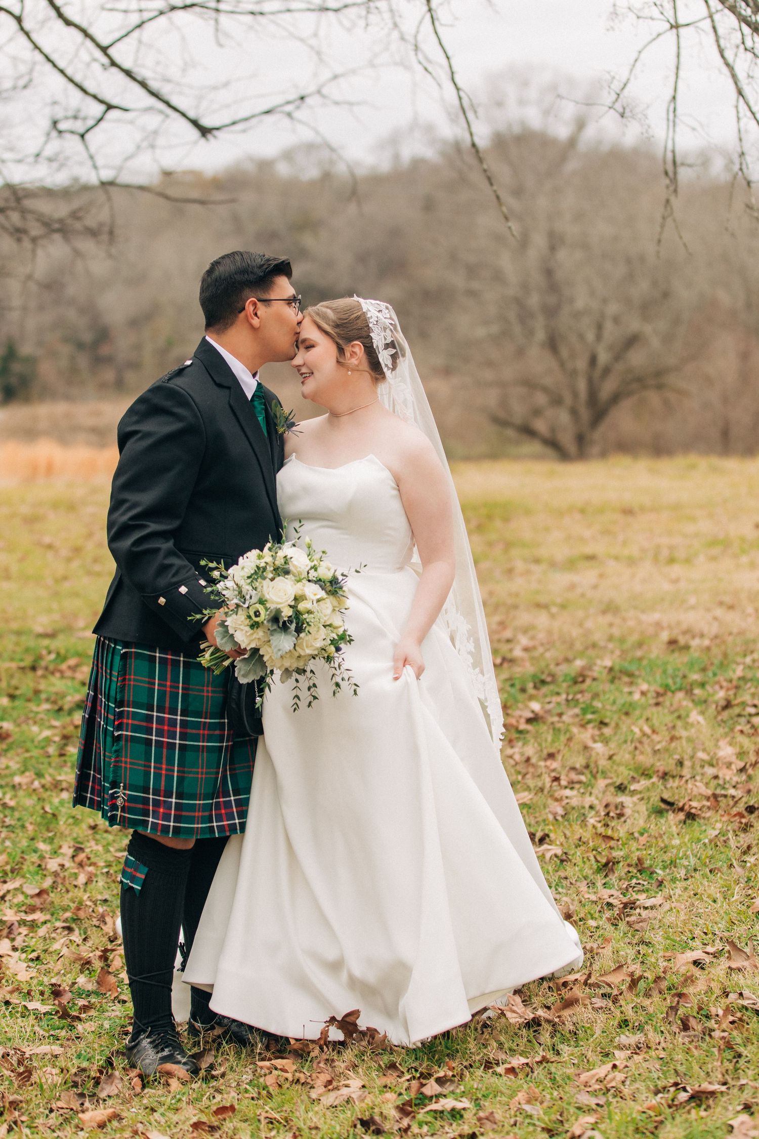 Newlywed couple embraces in rustic field setting with groom in traditional Scottish kilt and bride in white wedding gown.