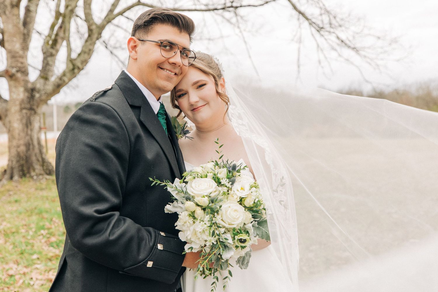 A newlywed couple embraces outdoors while holding a white floral bouquet on their winter wedding day.