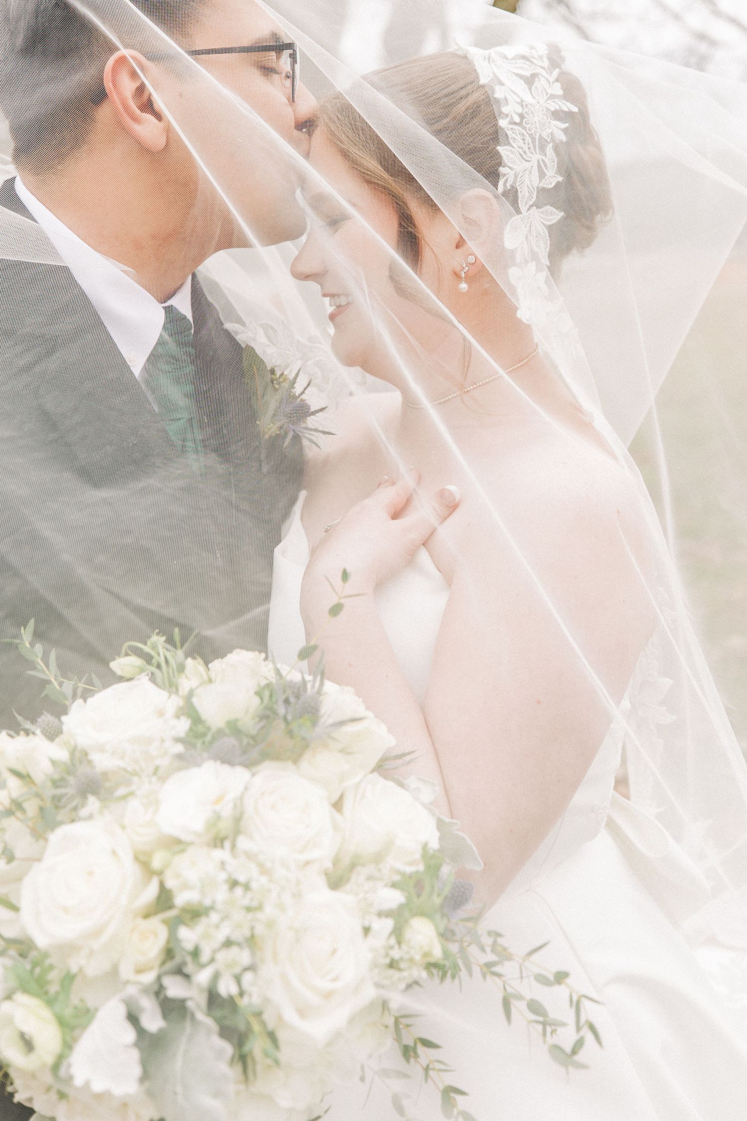 A romantic wedding moment captured through delicate lace veil with soft white floral bouquet in the background.