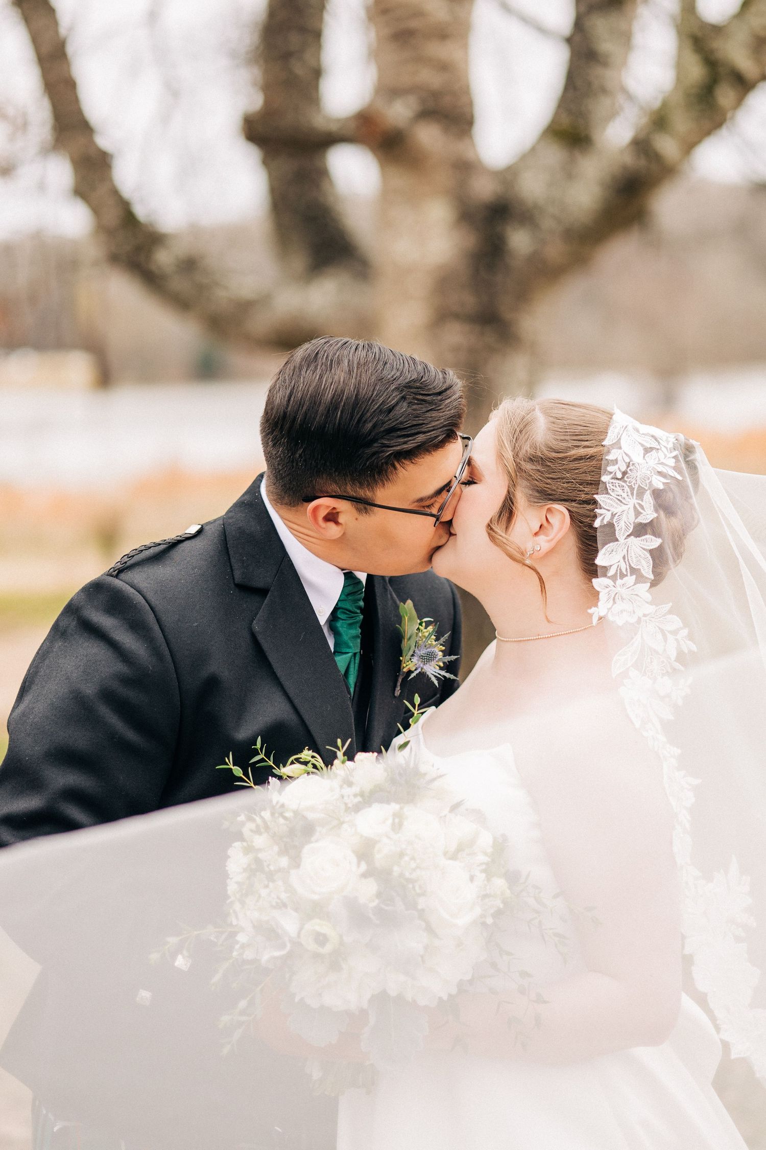 A bride and groom share a romantic kiss during their wedding ceremony while a delicate lace veil flows in the breeze.