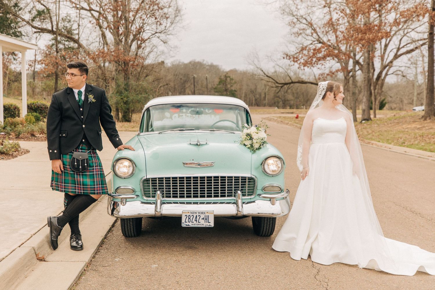 A turquoise vintage 1950s Chevrolet car sits between two people dressed in wedding attire on a scenic country road.