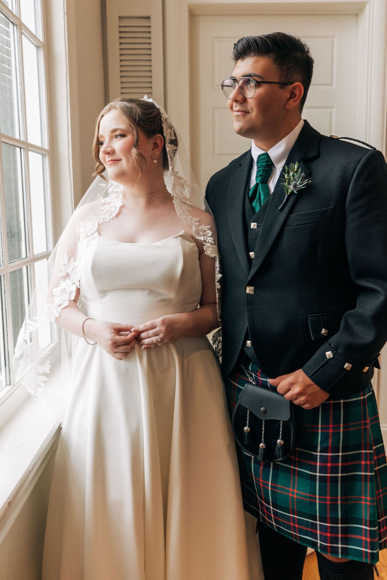 Newlyweds in formal attire pose by window, with bride in white gown and veil and groom wearing traditional Scottish kilt.