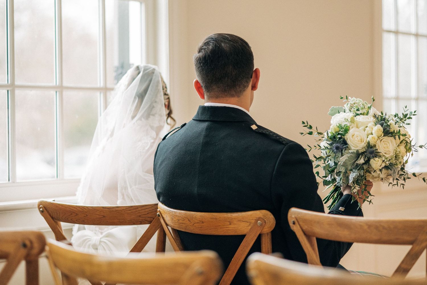 A couple seated at a wedding ceremony with wooden chairs and white floral bouquet visible from behind.