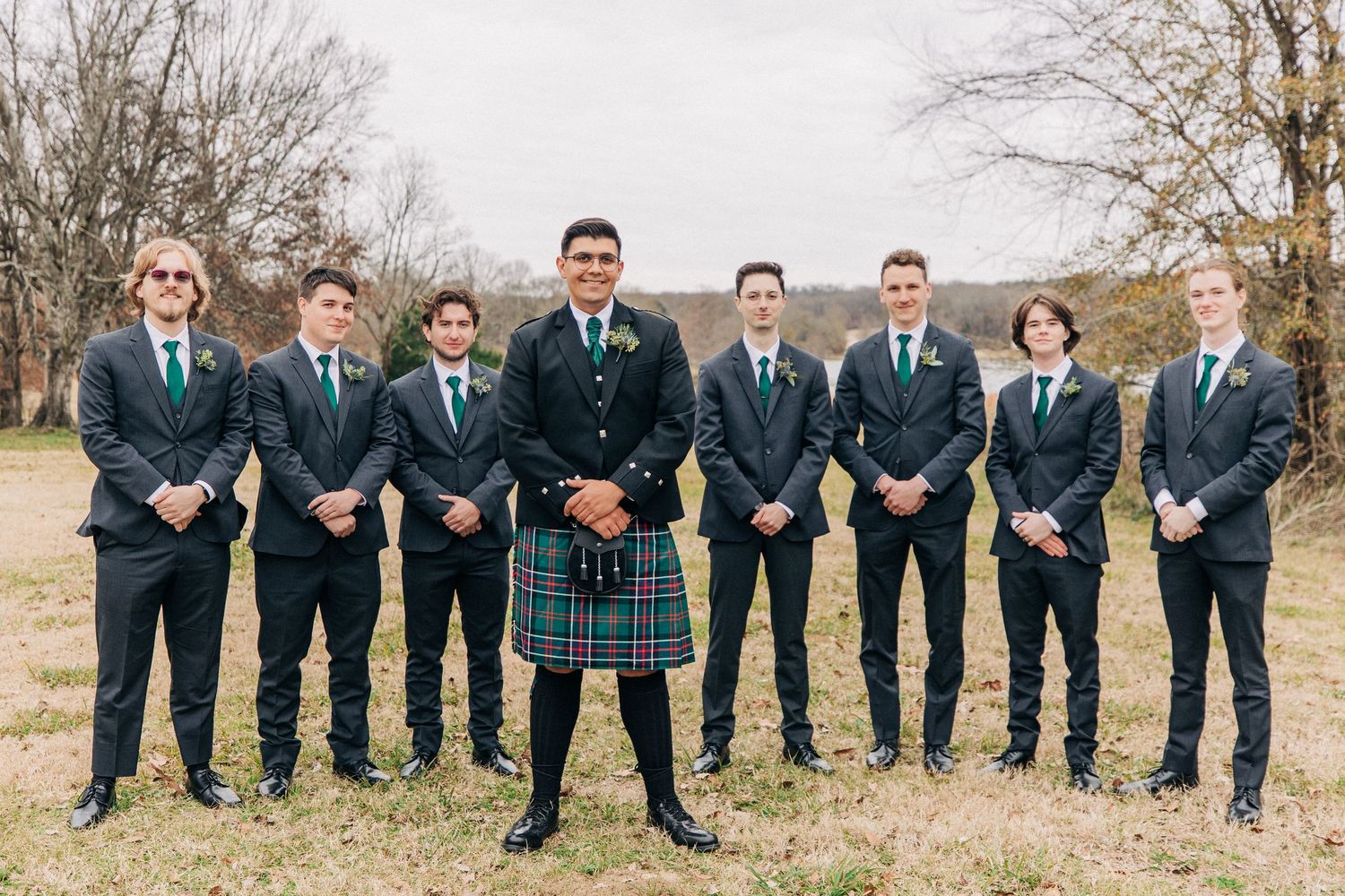 A group of groomsmen in matching navy suits stand outdoors, with one wearing a traditional Scottish kilt in the center.