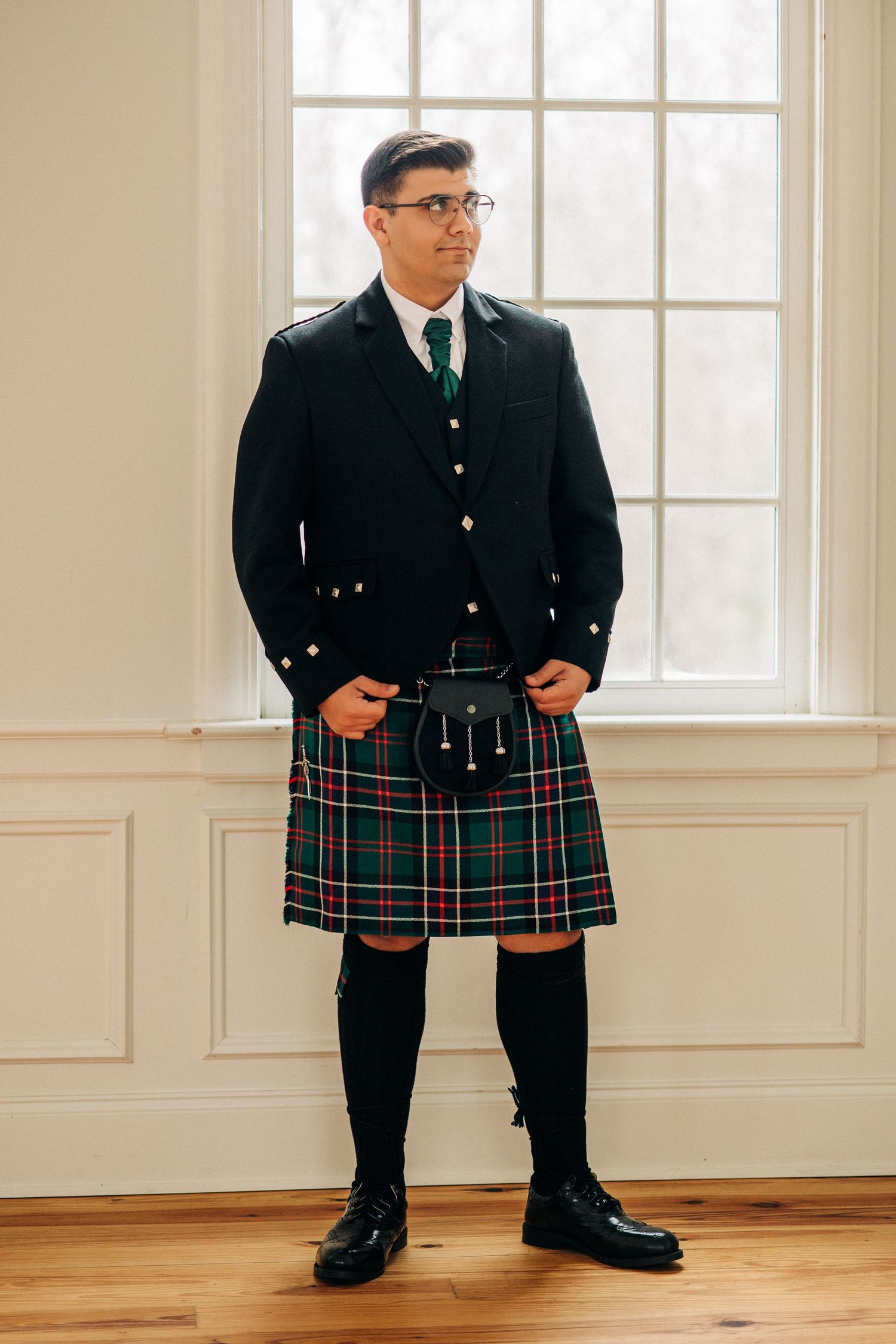 A person wearing formal Scottish attire with a green and red tartan kilt and black jacket stands by a window.