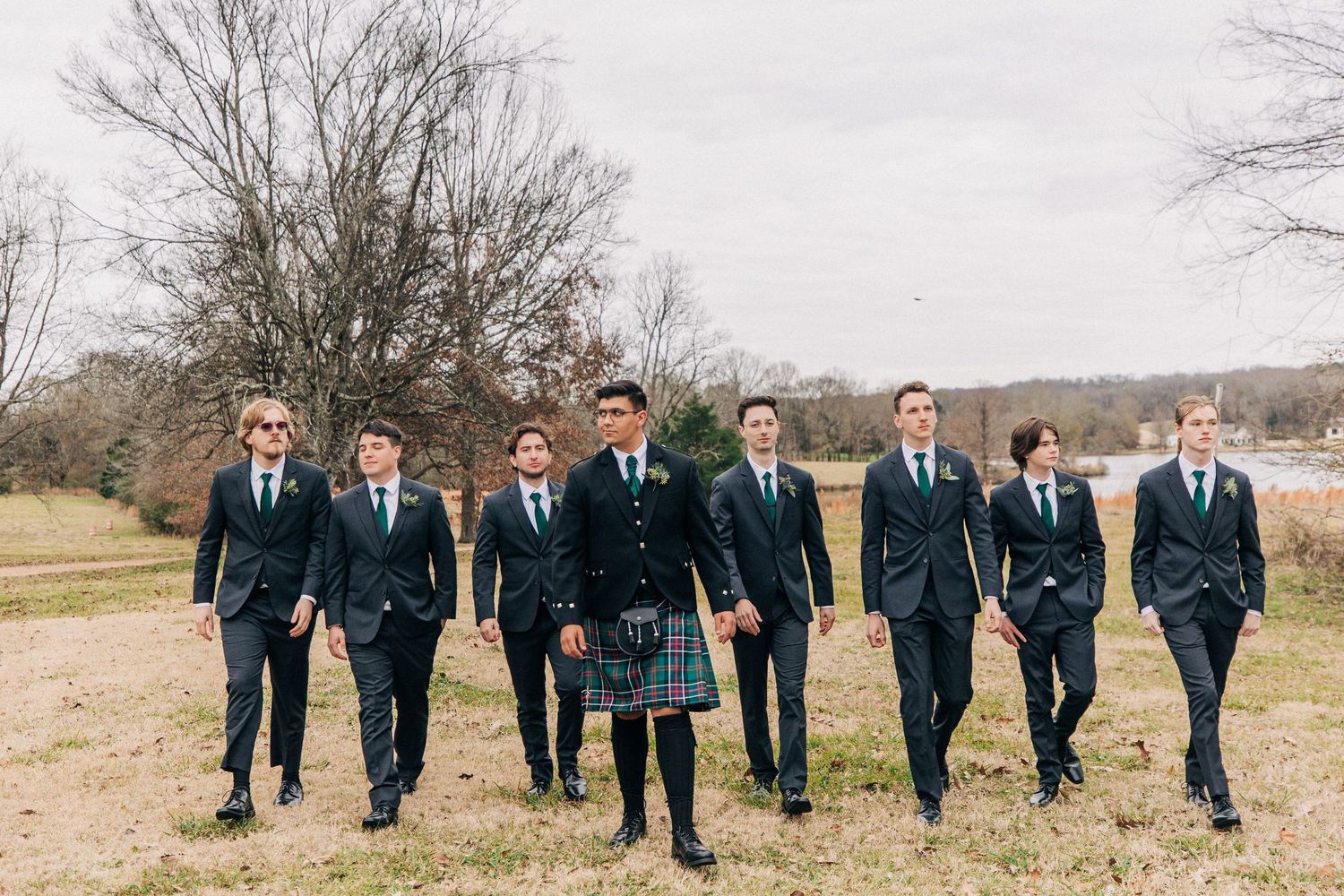 Group of young groomsmen in navy suits walking together across a grassy field on an overcast winter day.