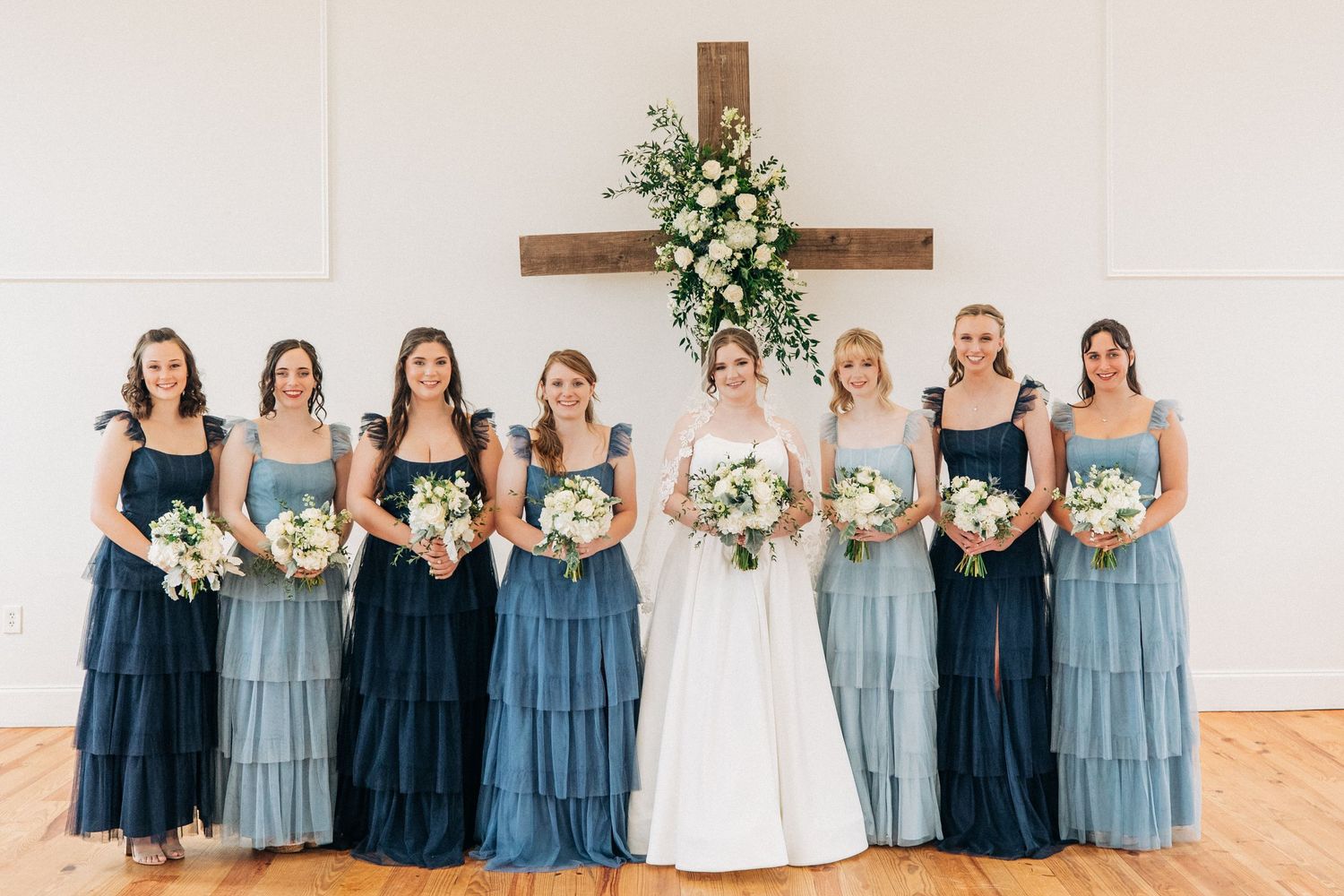 Bridesmaids in alternating navy and dusty blue dresses stand together holding white floral bouquets in church setting.