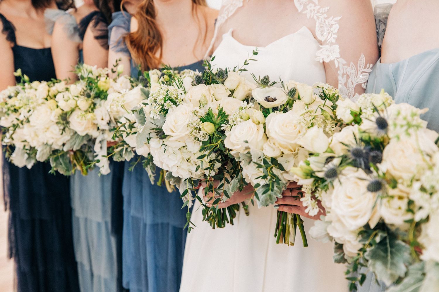 Row of white floral bouquets held against navy blue and light blue bridesmaid dresses at a wedding ceremony.