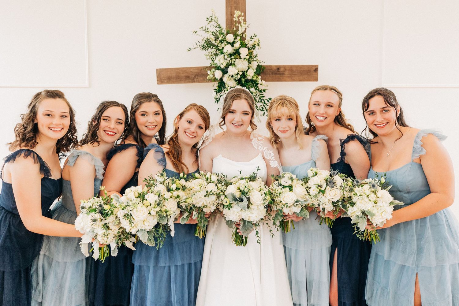 A bridal party wearing light blue bridesmaid dresses poses with their white floral bouquets in front of a wooden cross.