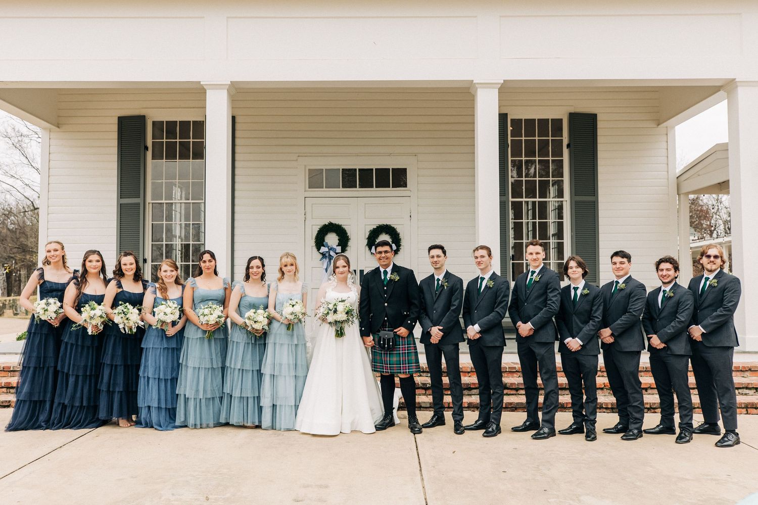Wedding party in formal attire poses on front porch of elegant white columned building with bridesmaids in blue dresses.