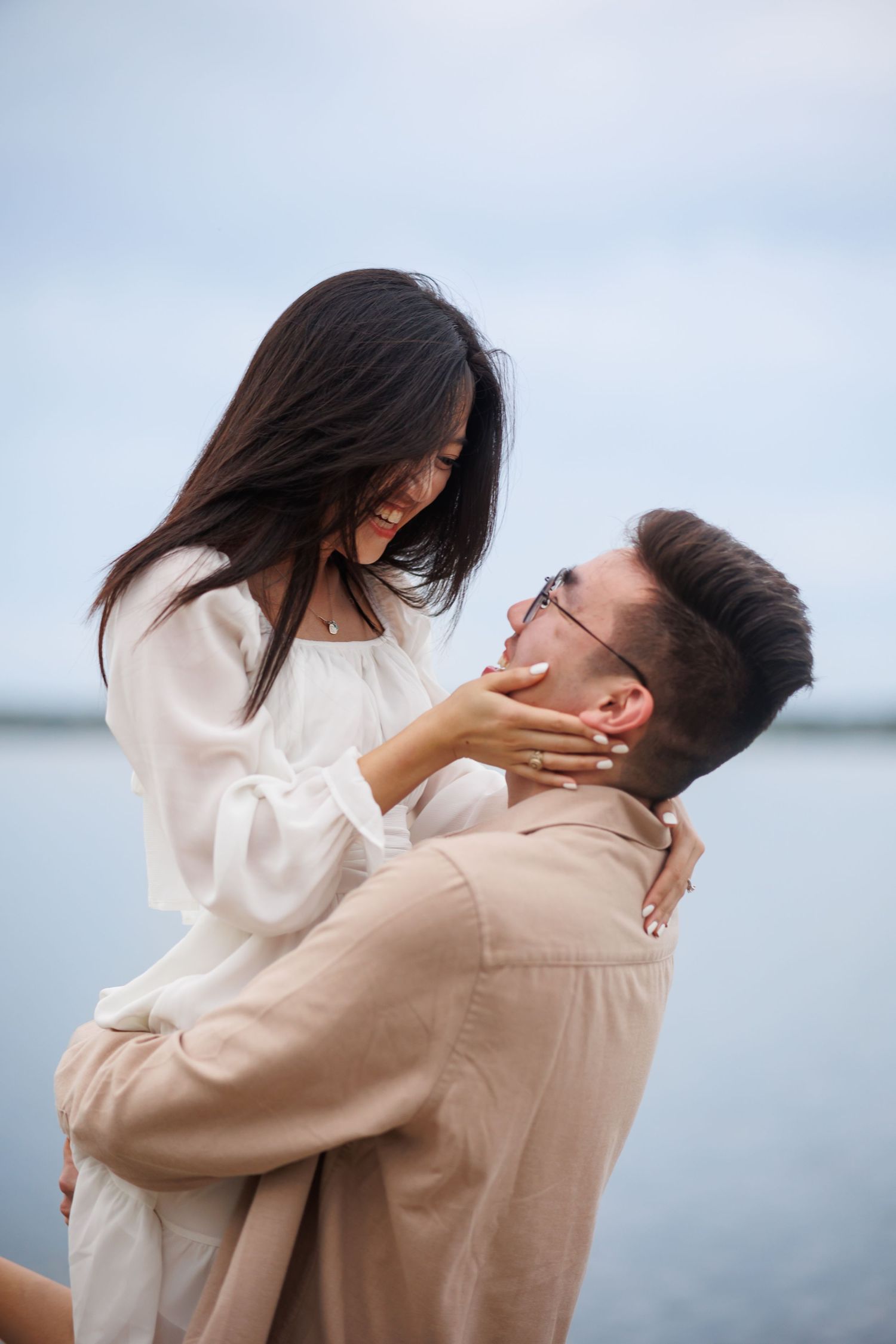 A romantic couple in neutral toned outfits embrace by the water during their summer Dallas engagement session.