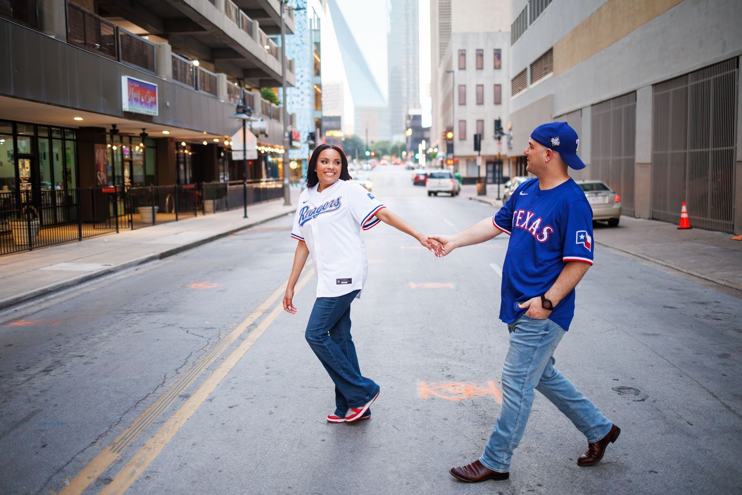 Couple dressed in matching baseball jerseys walk in a Dallas alley with city buildings in background during their engagement photoshoot