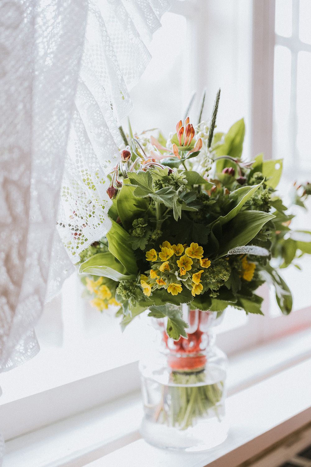 Bröllopsbukett i ett fönster med en vit spetsgardin, inspiration till lantligt bröllop. Brudbukett med gullviva, liljekonvalj och vårblommor