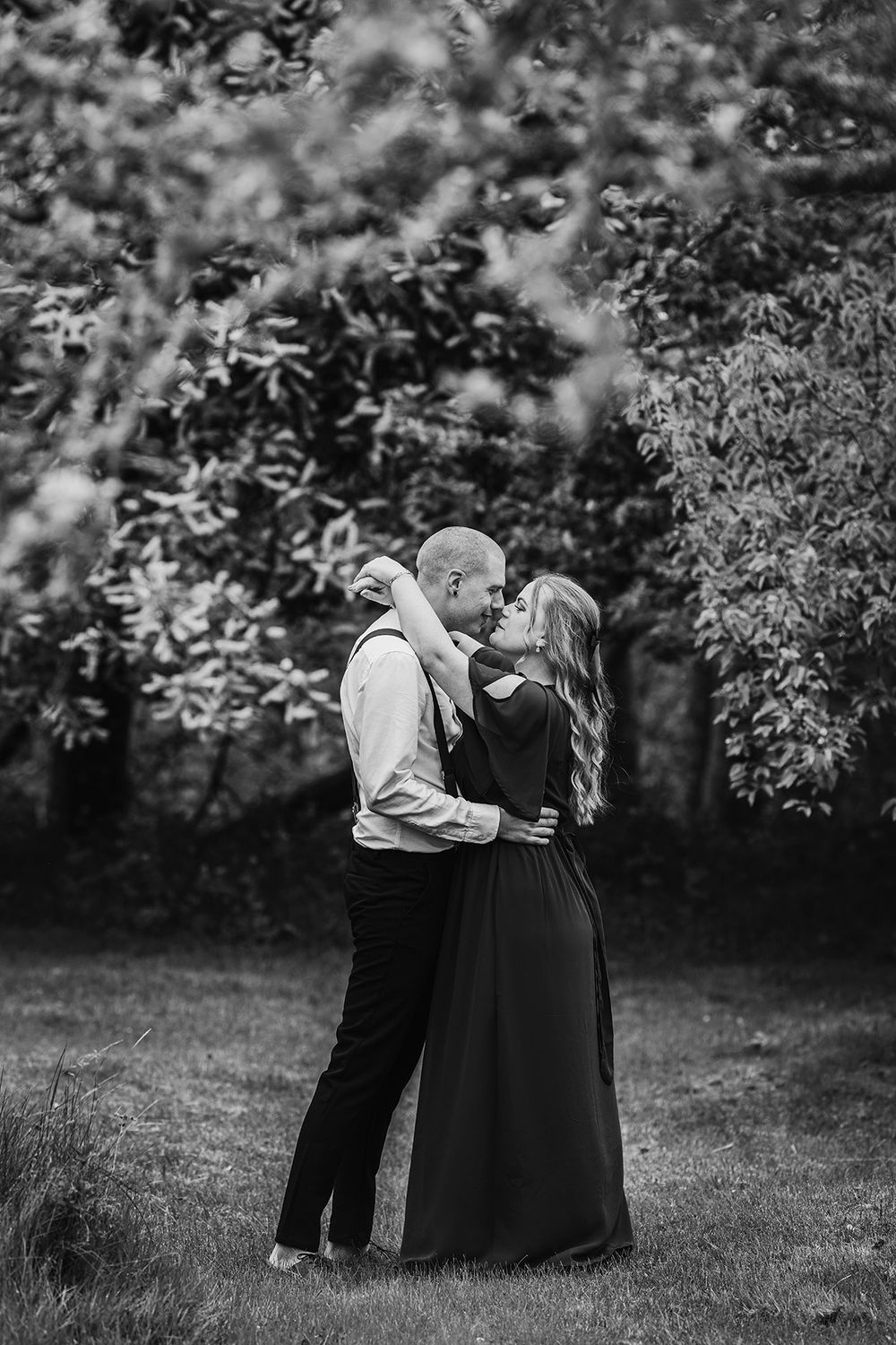 Bröllopsfoto i maj på brudpar som står i en trädgård med blommande äppelträd, de är vända mot varandra och ser varandra i ögonen.