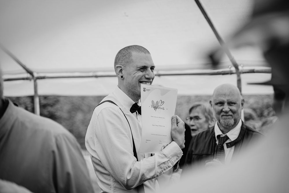 Brudgummen ler och håller upp ett papper som visar vigselbevis i sin hand. Bröllopsfest dokumenterad av Lets Bloom Media i Östergötland.