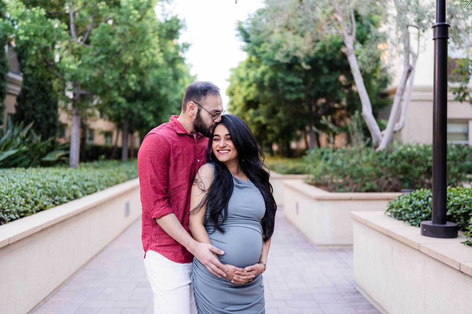 A tender maternity moment captured outdoors as couple stands together on a walkway surrounded by greenery.