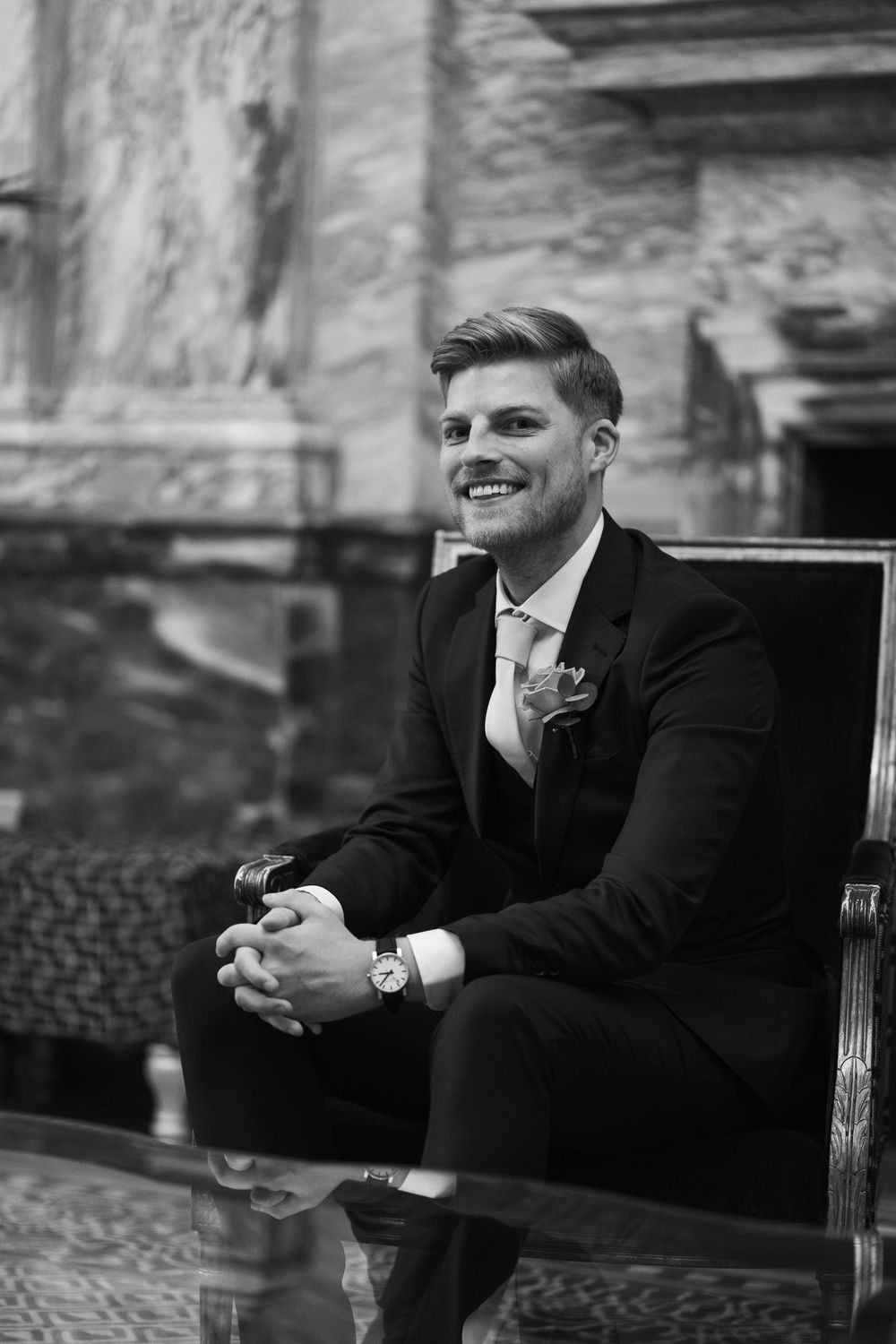 Groom in black suit smiles while sitting on elegant furniture in formal setting.