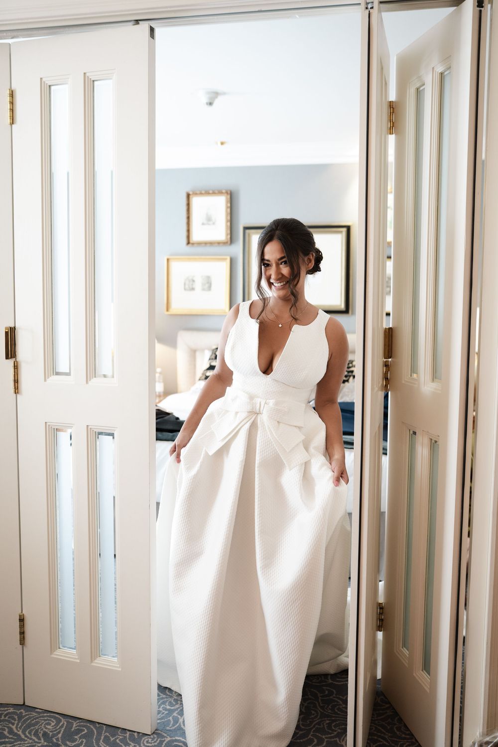 Bride in elegant white plunging neckline gown with bow detail stands in doorway.