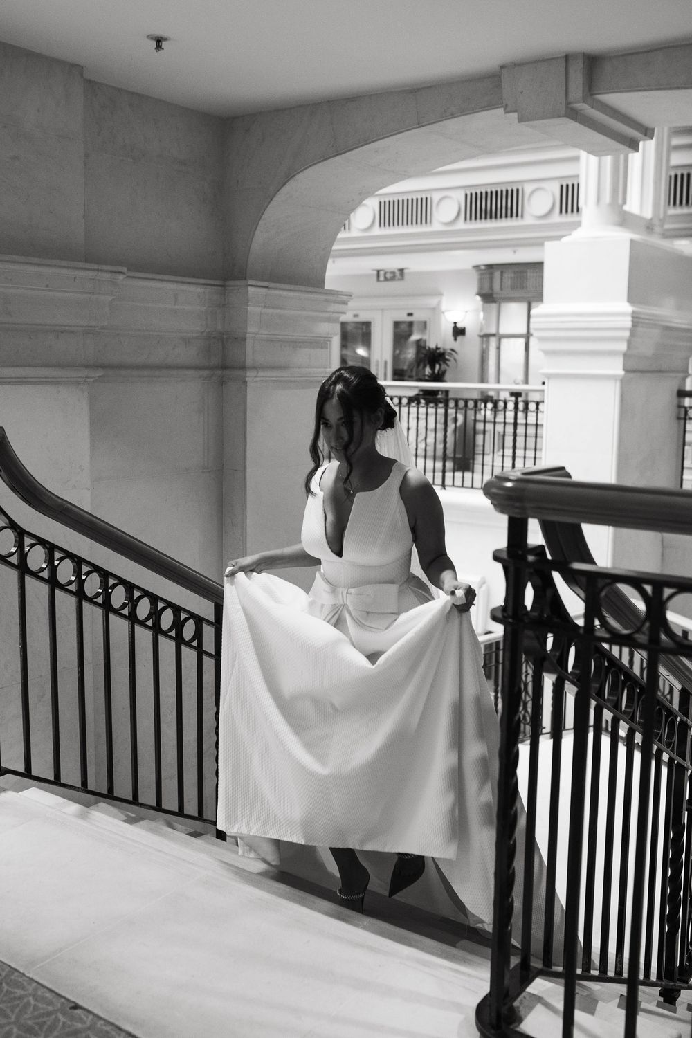 Bride holding white wedding dress on elegant staircase with iron railings.