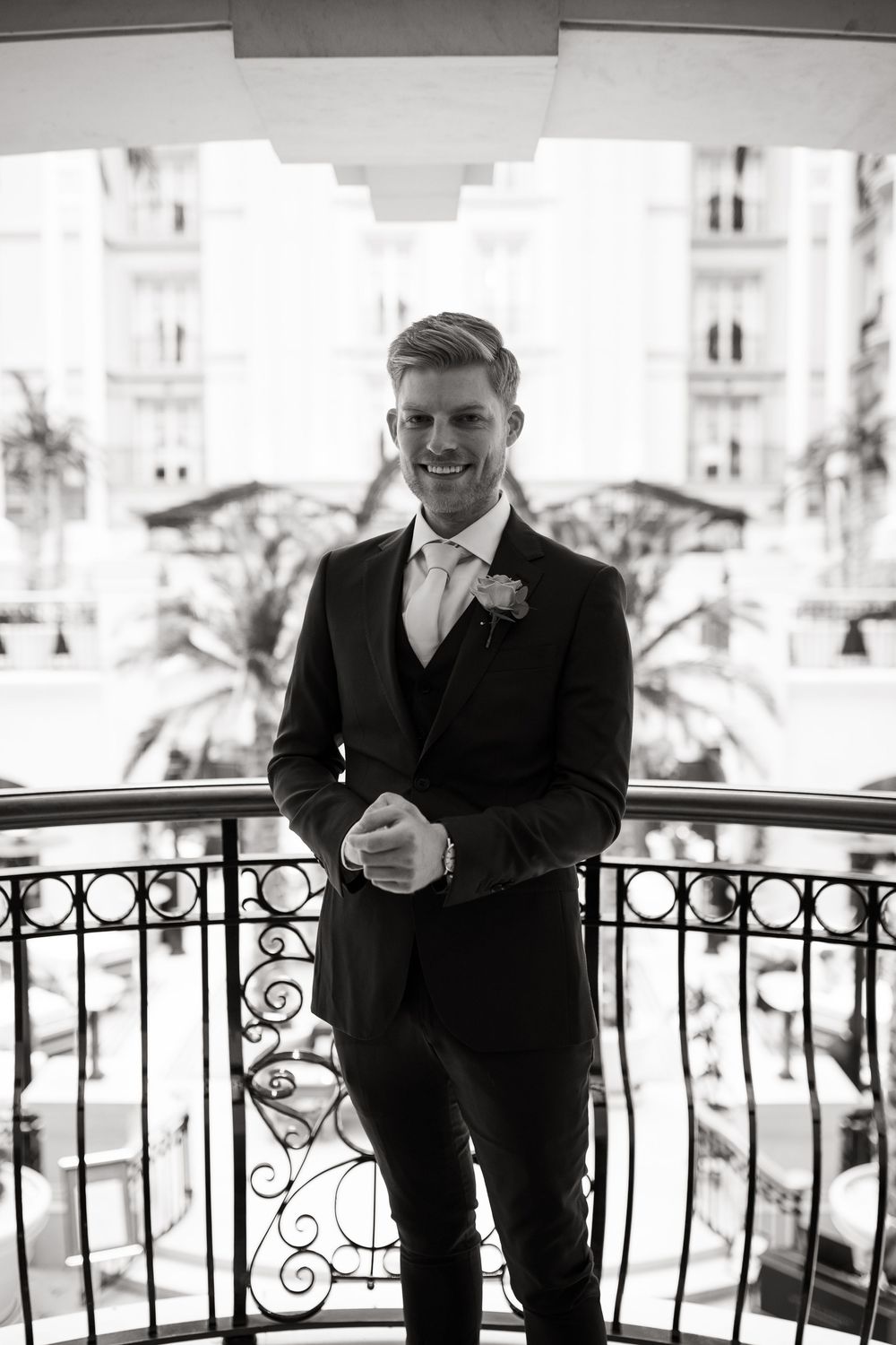 Groom in black suit poses on ornate balcony in black and white photo.