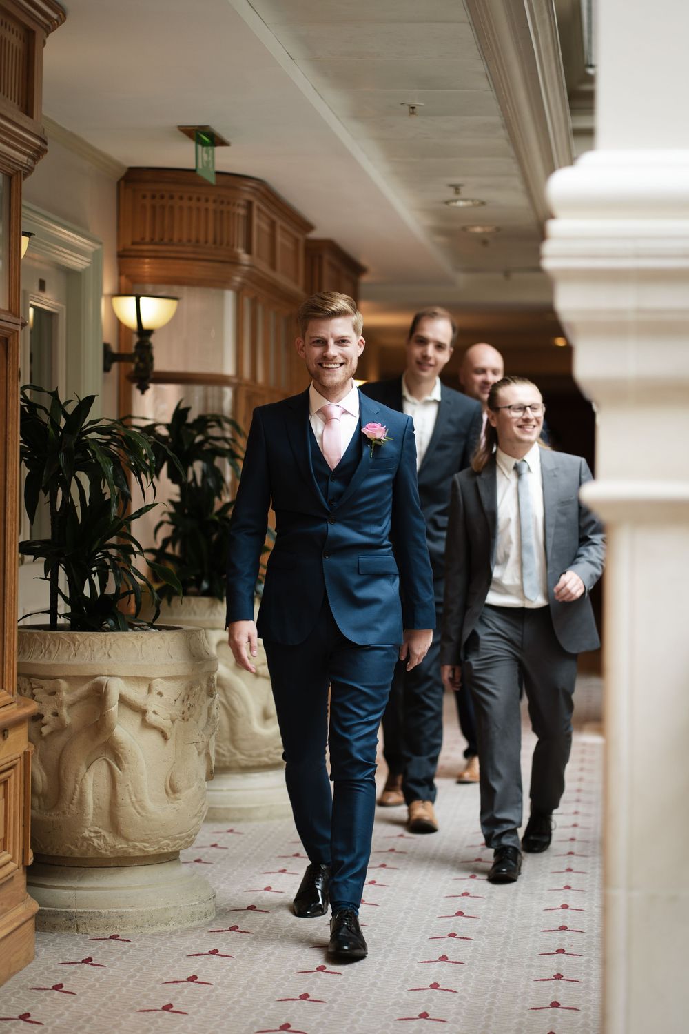 Groom in navy suit walks confidently through elegant hotel corridor with groomsmen.