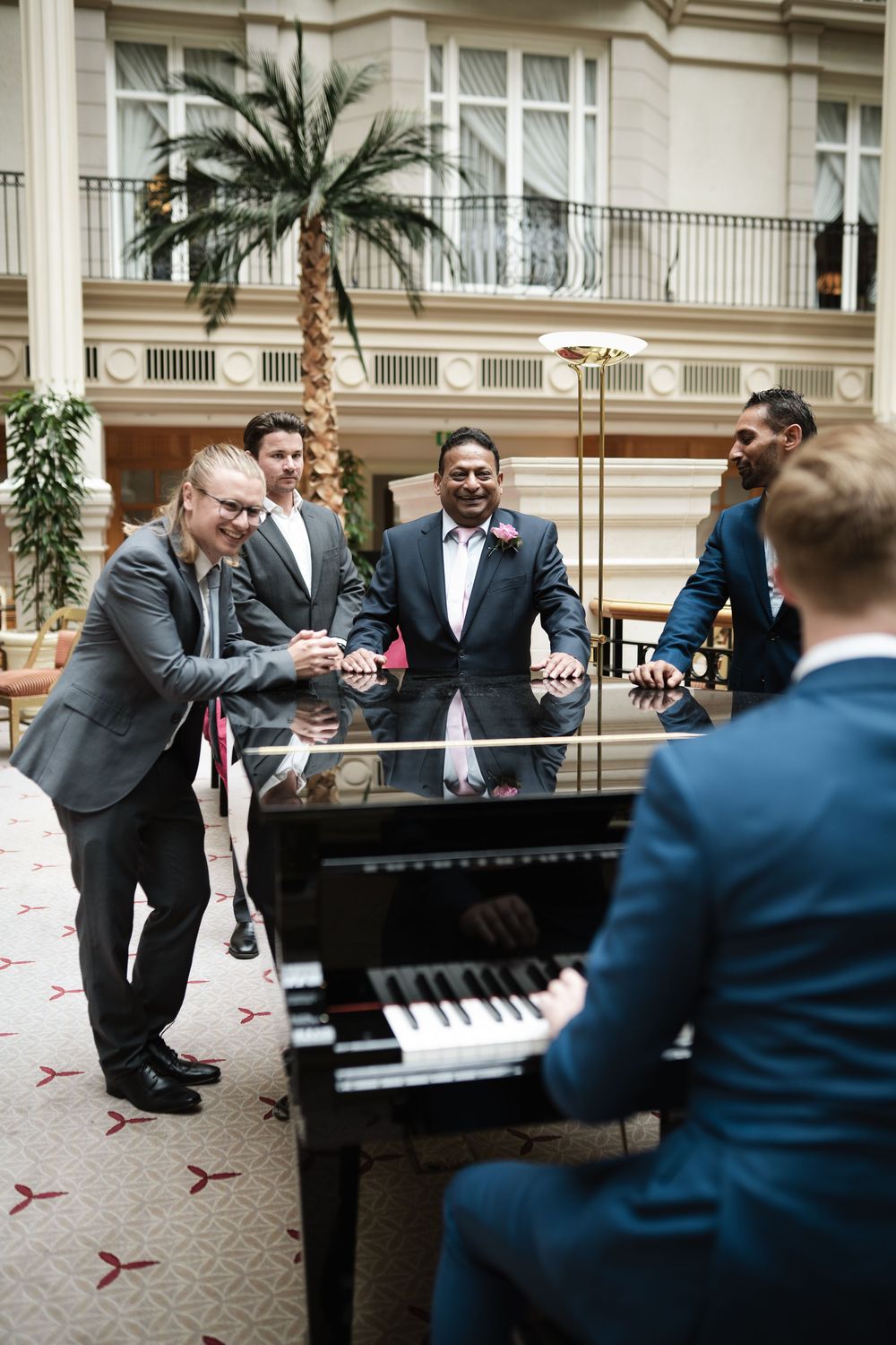 Group of men in suits gather around grand piano during wedding celebration.