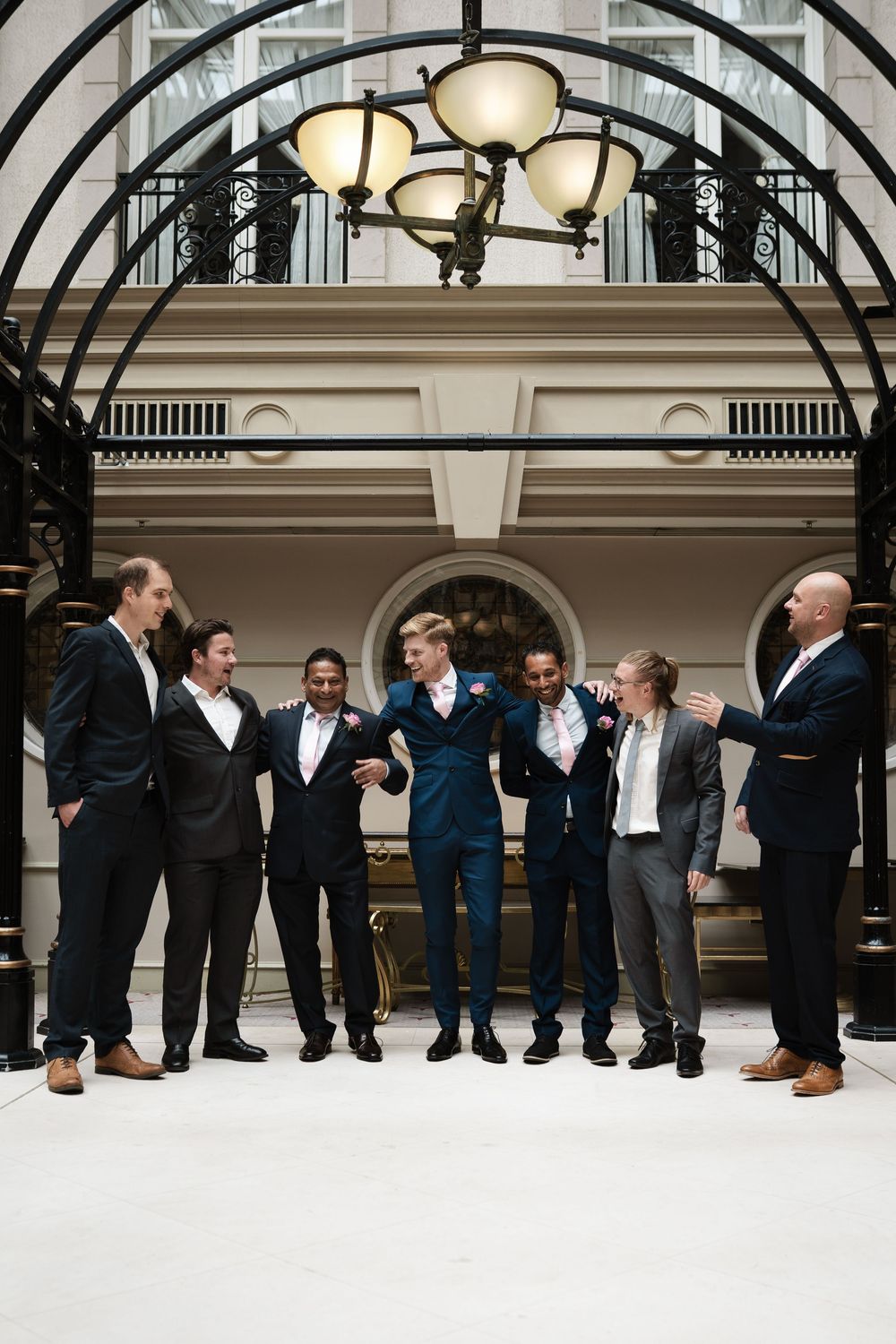 Groomsmen in dark suits pose under elegant arched ceiling with chandelier.