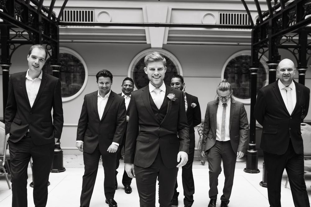 Black and white photo of groomsmen walking in formation under ornate architecture.