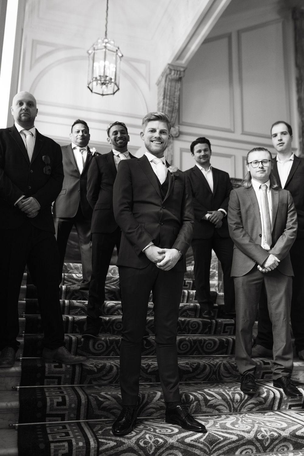 Groomsmen in formal attire pose on an elegant staircase with ornate carpeting and classic lighting.