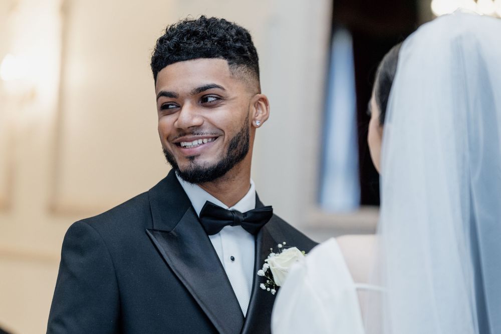 Groom wearing black tuxedo and bowtie smiles warmly during wedding ceremony.