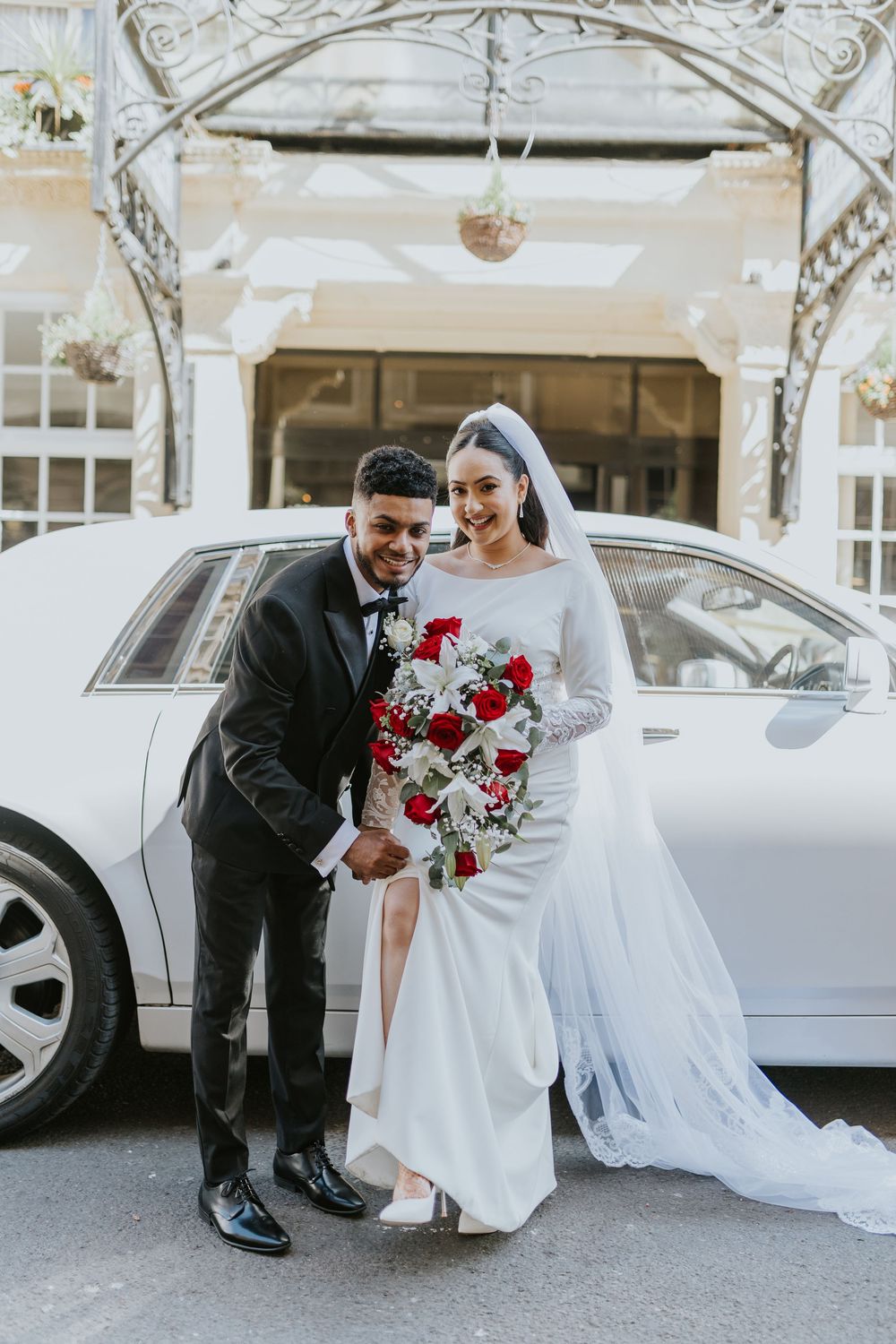 A happy couple poses with a stunning red rose bouquet in front of a luxury white car on their wedding day.