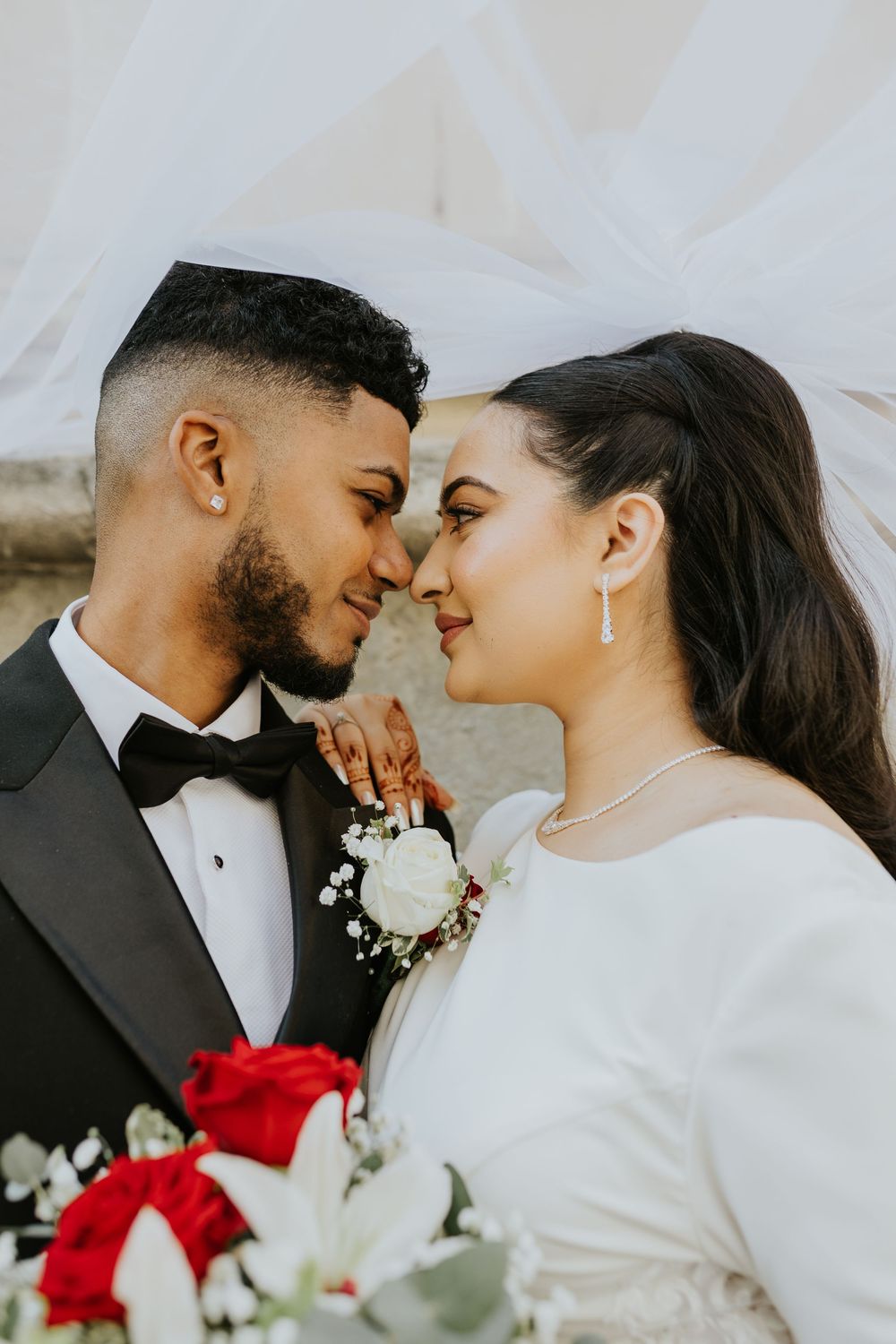 Romantic wedding portrait featuring a tender moment between newlyweds with red and white bouquet against light background.