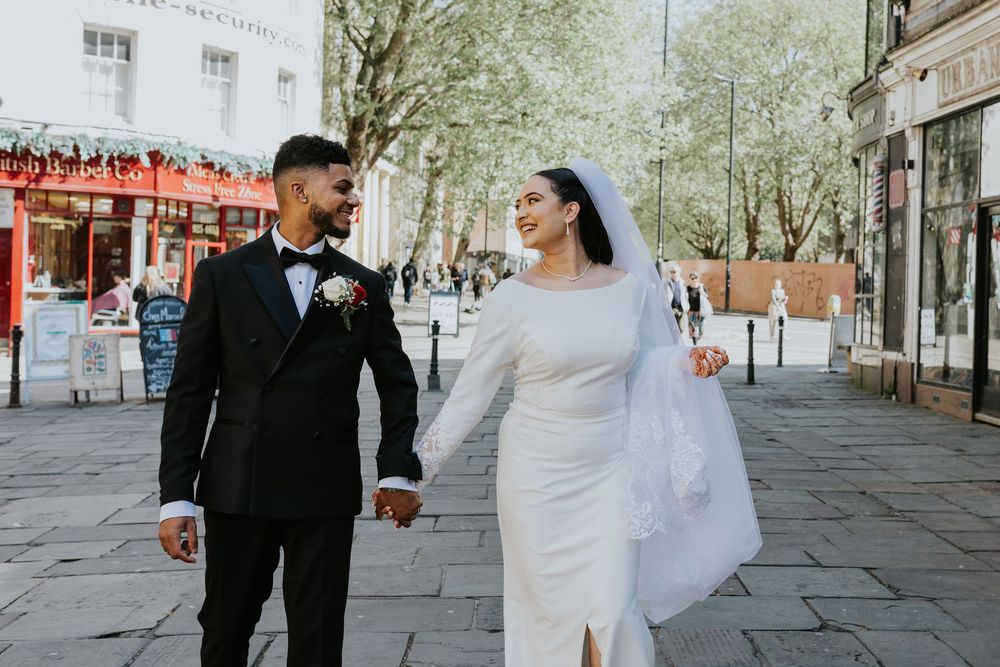 A newlywed couple walking hand in hand on a cobblestone street with storefronts and spring trees in the background.