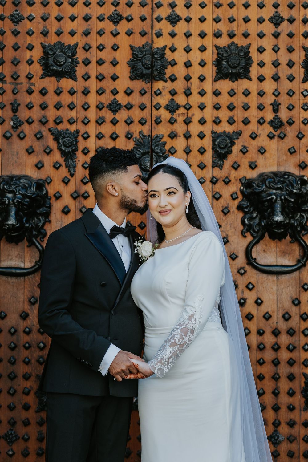 Elegant bride and groom pose against ornate wooden door with decorative lion head door knockers.