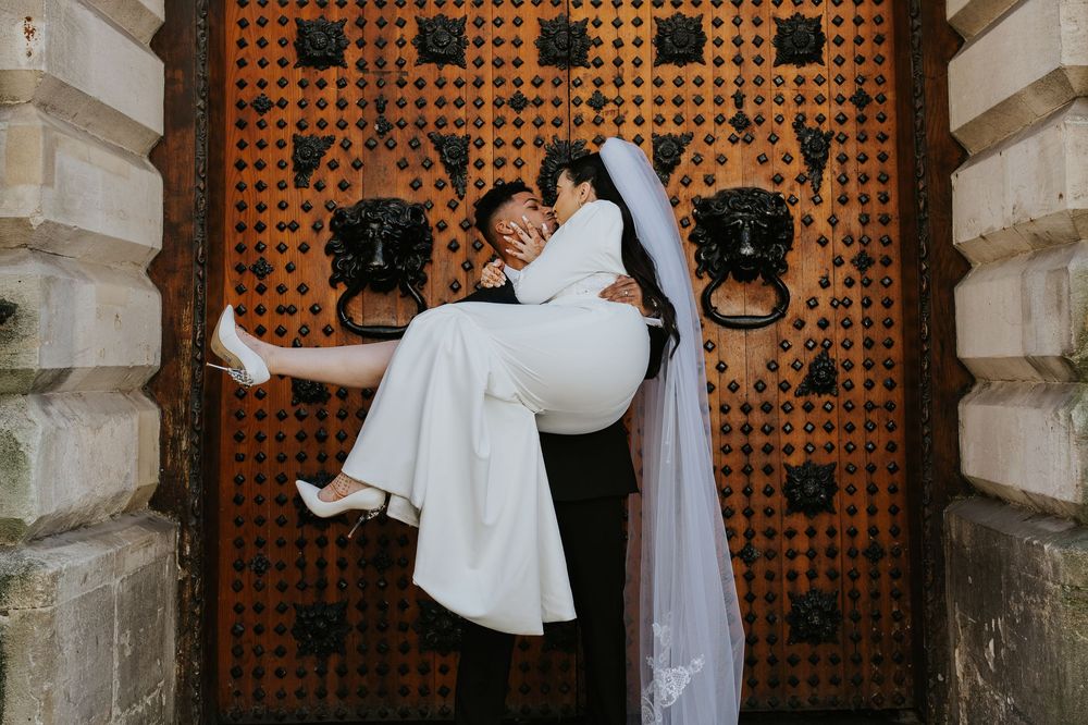 A joyful couple wearing wedding attire pose in front of an ornate wooden door with decorative black iron hardware.