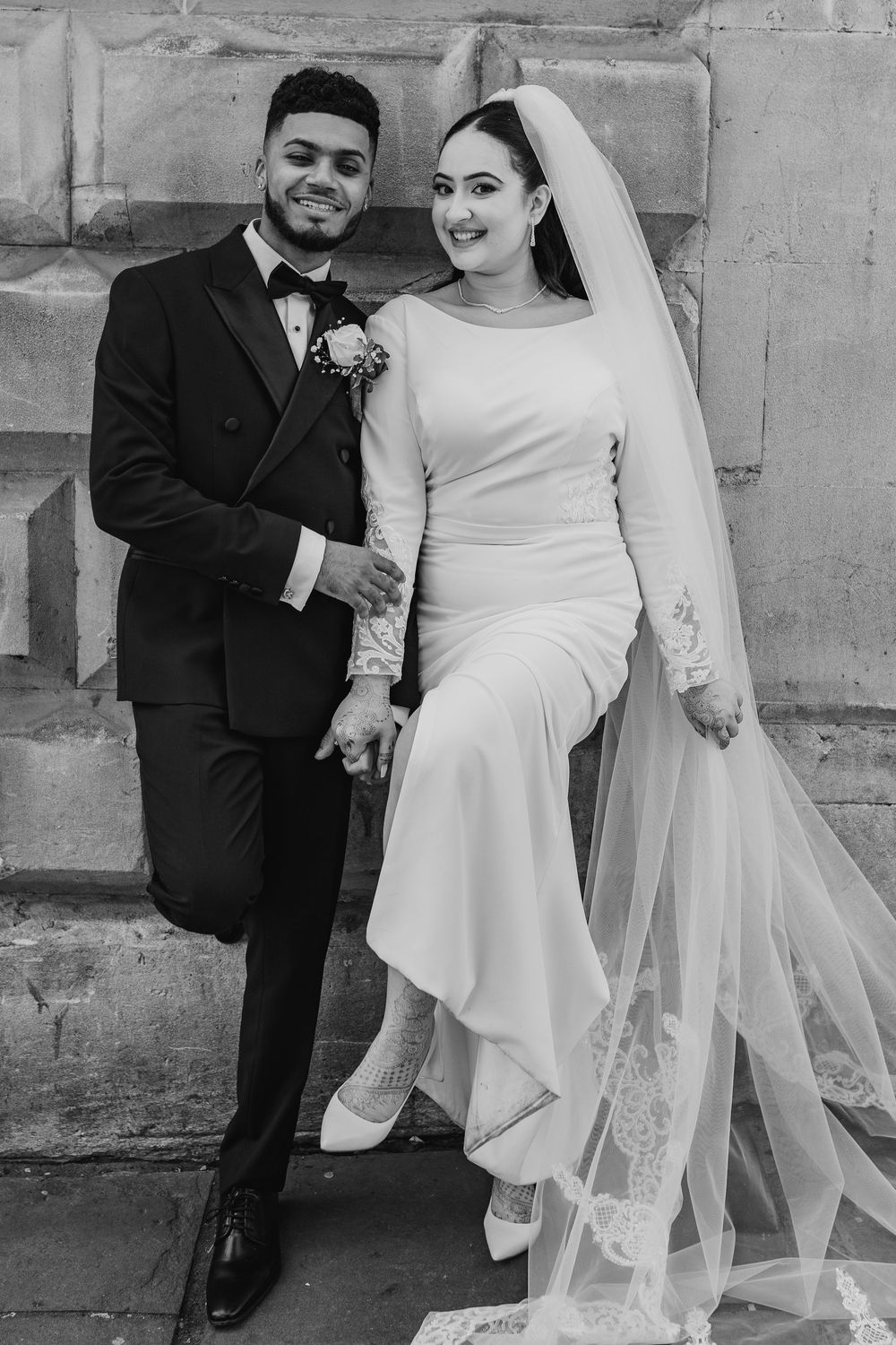 A black and white wedding portrait of a couple posing against a stone wall, the bride in an elegant white gown and veil.