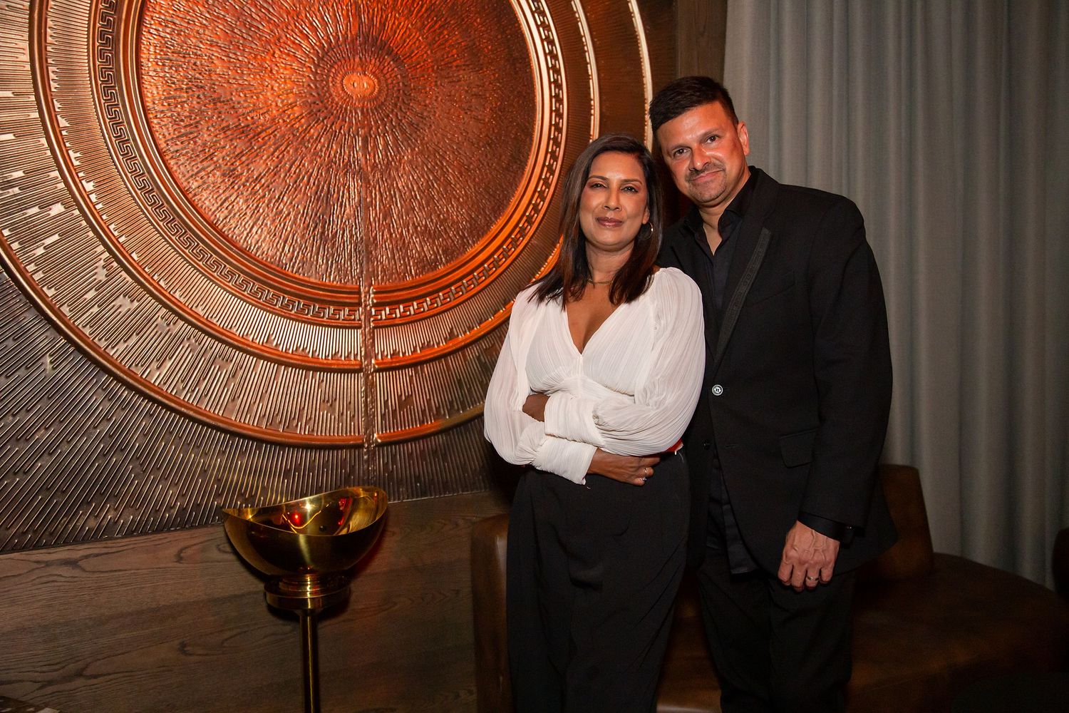Two people pose together at an elegant event venue with a large decorative copper wall medallion behind them.