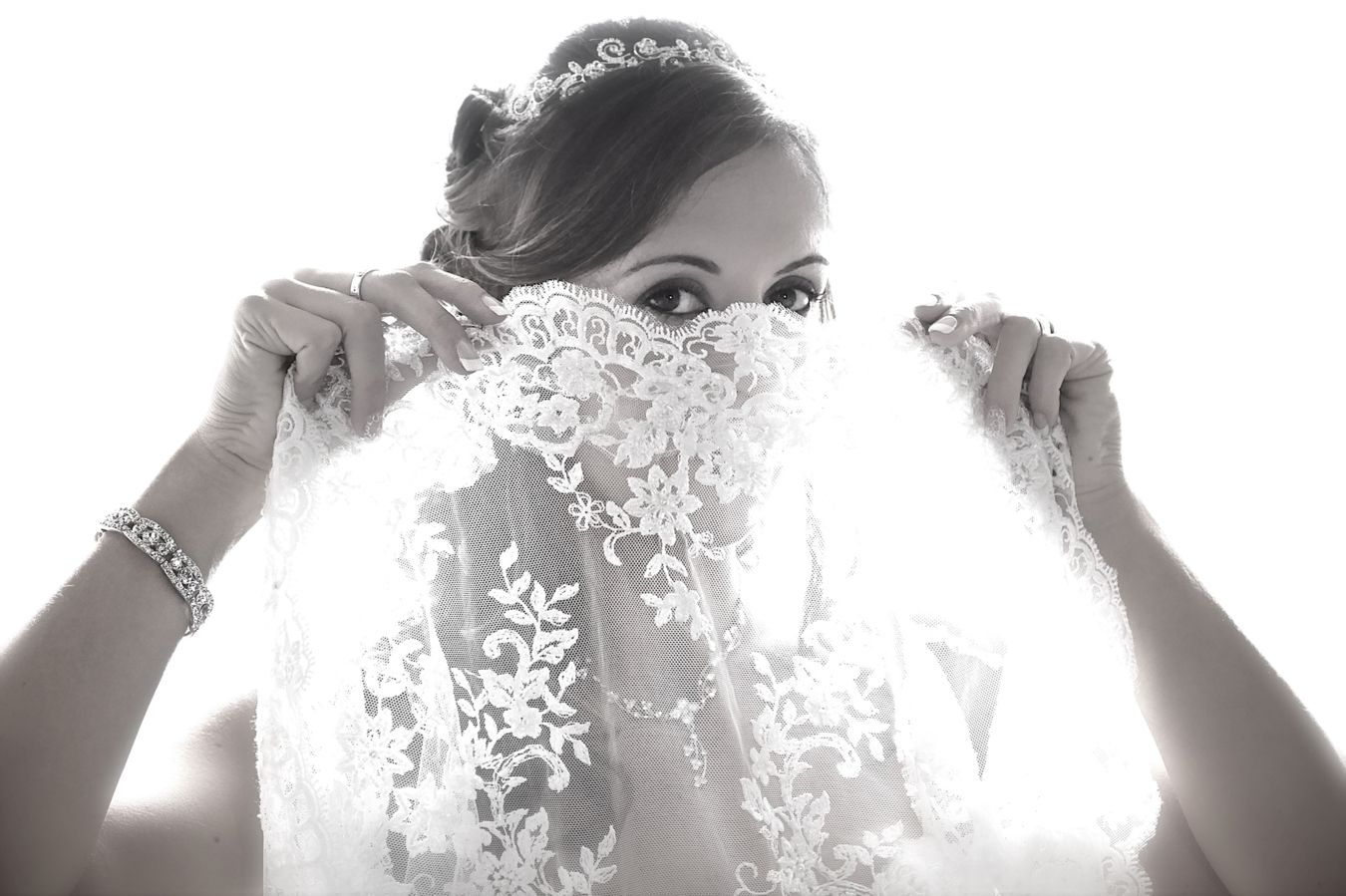A black and white photo shows a bride holding up delicate lace veil in front of bright backlighting.