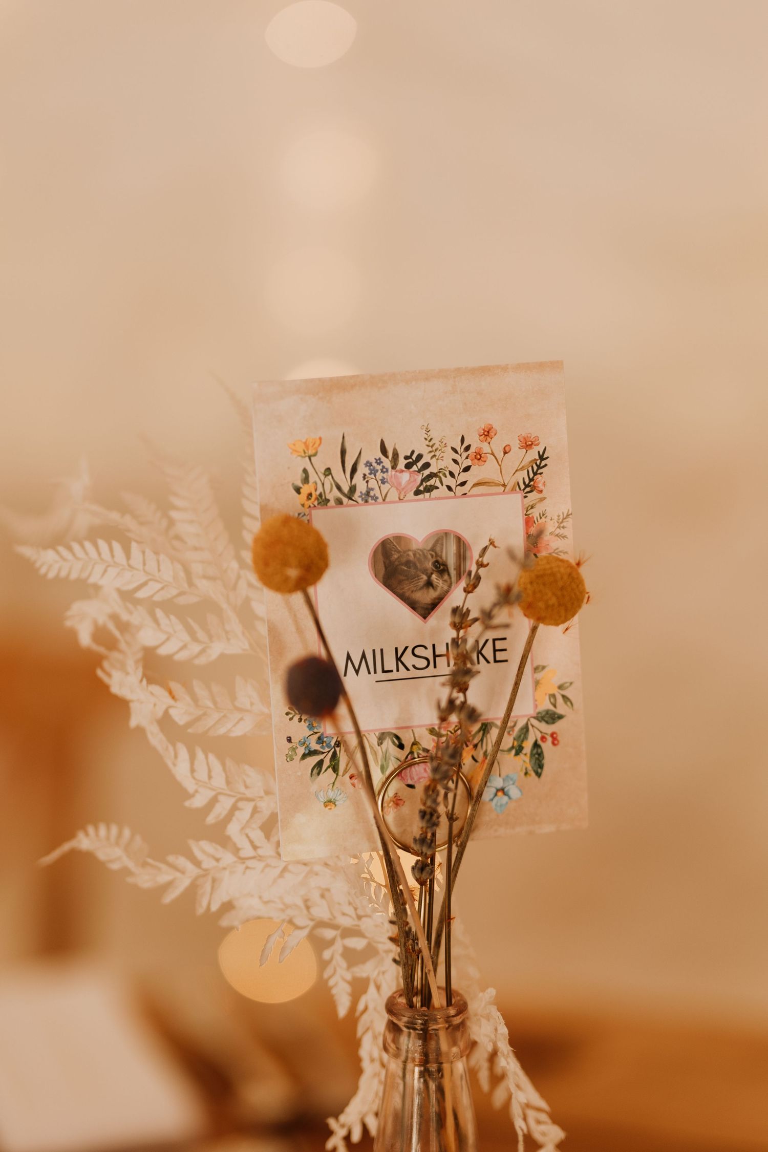 A vintage-style milkshake card with dried flowers and pampas grass in a vase against a warm beige background.