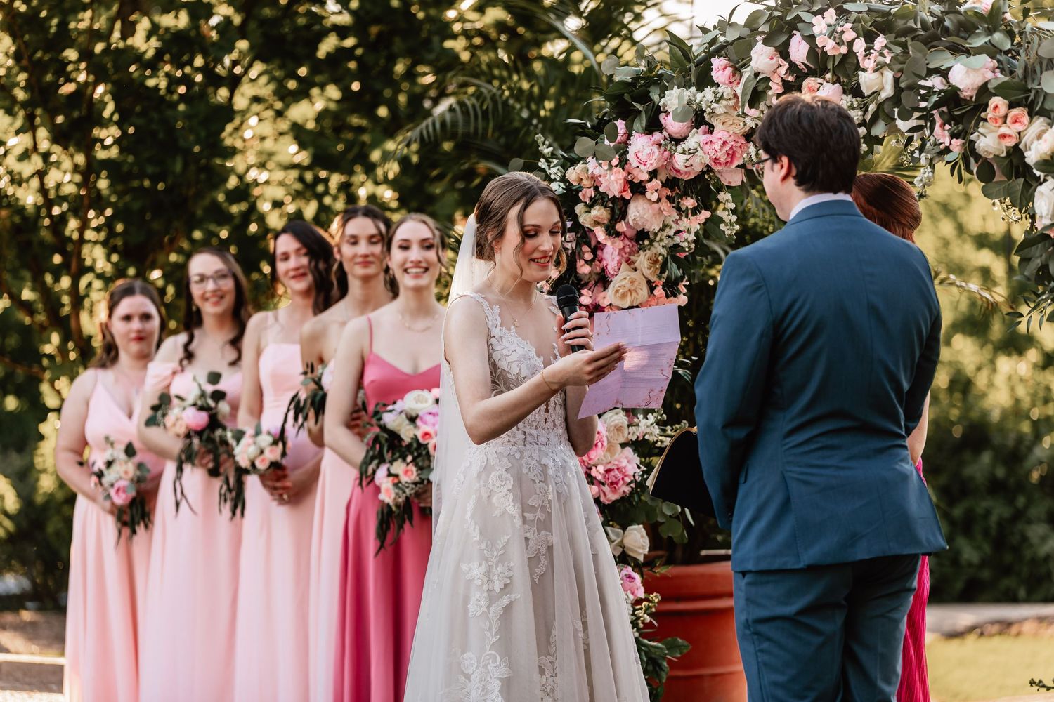 Bride in white lace gown exchanges vows with groom while bridesmaids in pink dresses watch during outdoor ceremony.