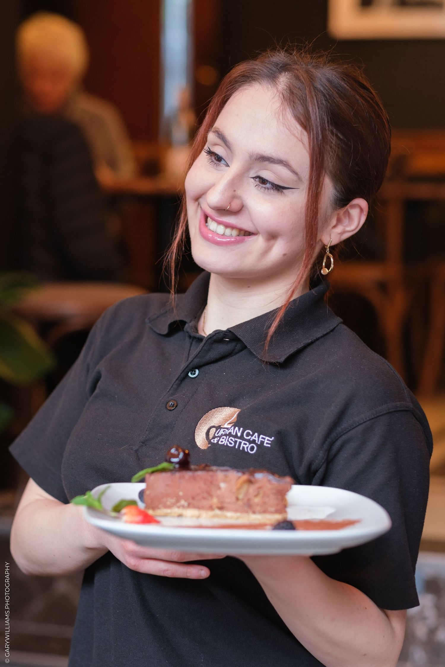 Server in black polo shirt presents a plated meal with a warm smile.