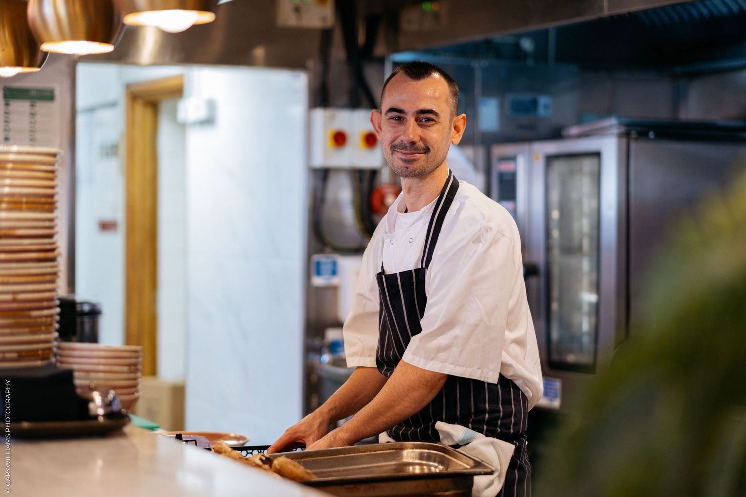 Restaurant staff member in white uniform and black apron preparing food at counter.
